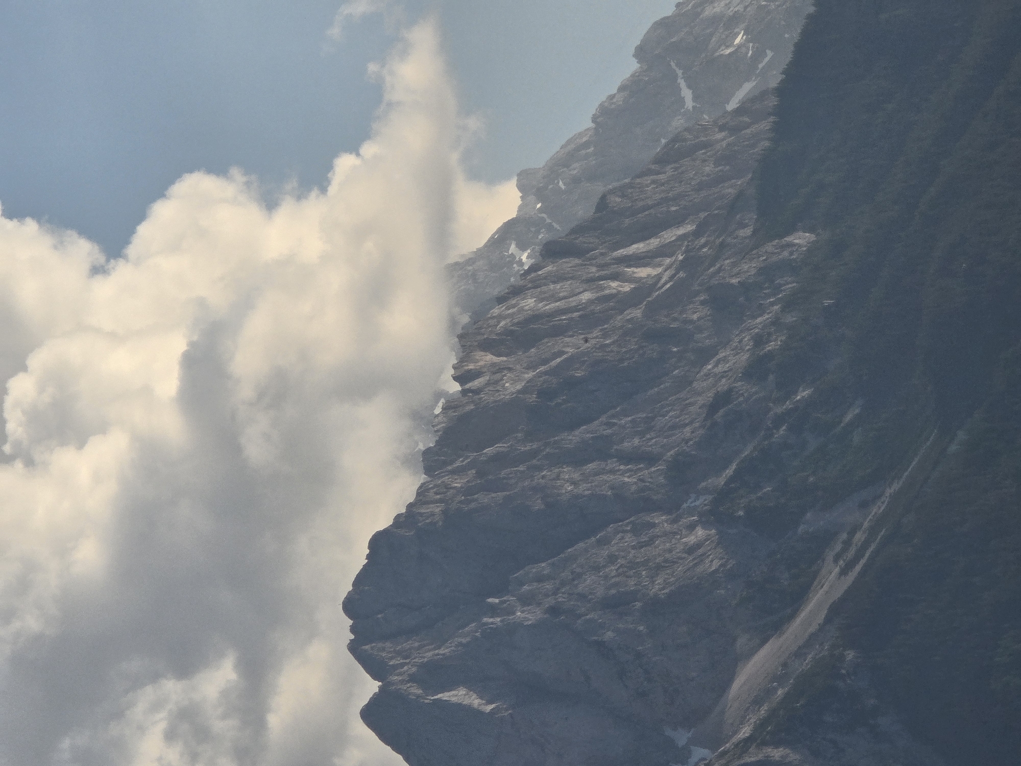 The image captures a dramatic mountain landscape in Garmisch-Partenkirchen, Germany. The scene is dominated by the imposing, rocky face of a mountain, which fills the right two-thirds of the frame. The mountain's surface is textured with ridges and crevices, suggesting a rugged and ancient formation. Patches of snow are visible in some of the depressions, hinting at cooler temperatures or recent snowfall.
To the left of the mountain, fluffy white clouds are gathering, some of them partially obscuring the mountain's edge, creating a sense of depth and atmosphere. Beyond the clouds, the sky is a clear, light blue, indicating fair weather. The lighting suggests it is daytime, likely late morning or early afternoon, given the brightness of the sky and the way light falls on the mountain.
The overall mood is one of majestic natural beauty, with a touch of mystery introduced by the swirling clouds. There are no people or man-made objects clearly visible in the frame, focusing the viewer's attention entirely on the grandeur of the alpine scenery. The image evokes a sense of awe and the vastness of nature.