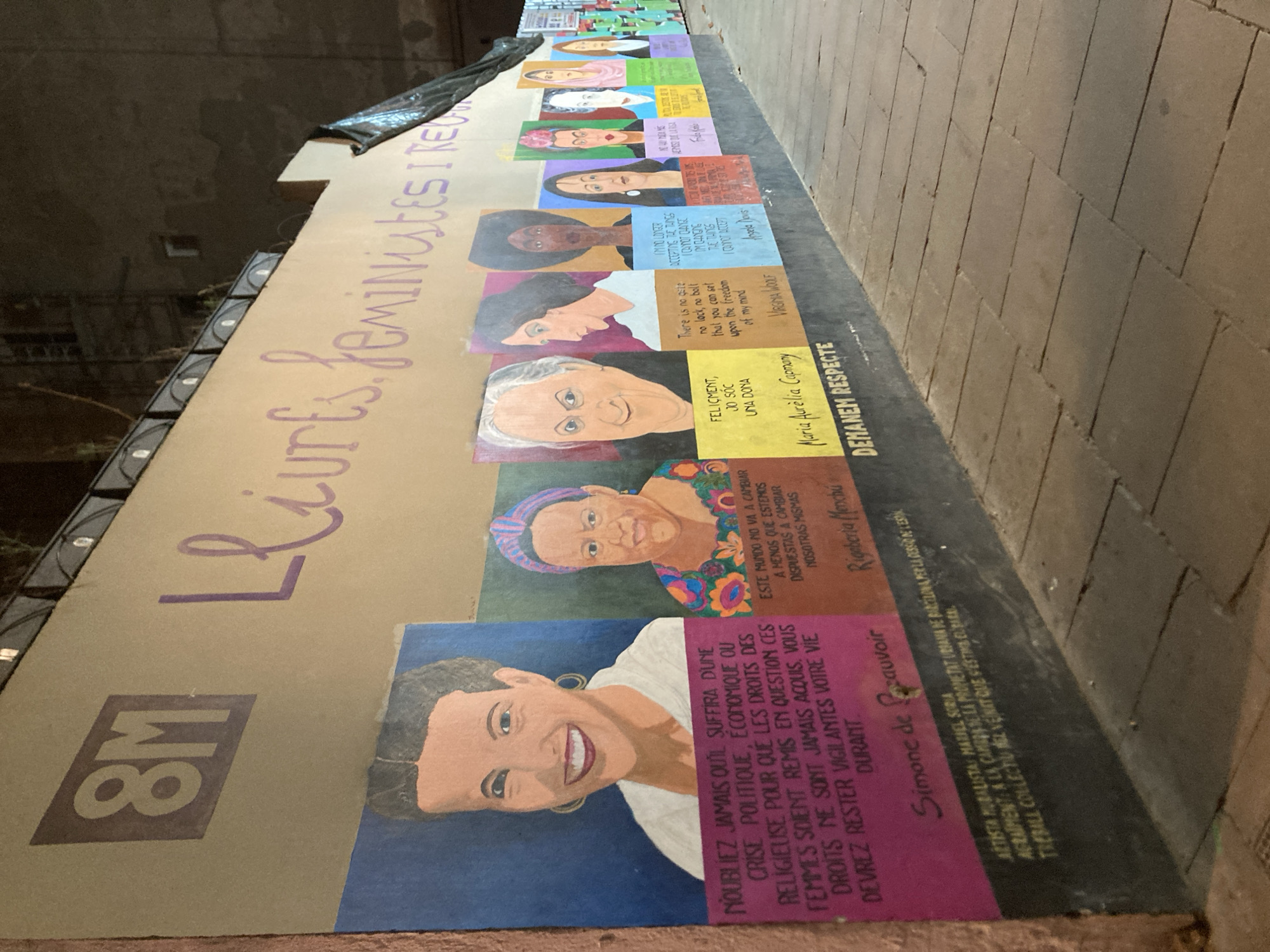 This image shows a long mural on a wall in Barcelona, Spain. The mural consists of portraits of women, likely feminists, with quotes attributed to them. The text "8M Lliures, feministes i rebel" is also written on the mural. The mural includes the following women: Simone de Beauvoir, Rigoberta Menchú, Maria Aurèlia Capmany, Virginia Woolf, Angela Davis, Frida Kahlo. The quote from Simone de Beauvoir reads, "N'oubliez jamais qu'il suffira d'une crise politique, économique ou religieuse pour que les droits des femmes soient remis en question. Ces droits ne sont jamais acquis, vous devrez rester vigilantes votre vie durant". Below Simone de Beauvoir it is written in small print: Artista Muralista Mariel Soria
Agraïment: A la Camio Ase La Propietat Urbana De Barcelona per la cesio de l'Espai: Treball collectin vel vent que s'està cuxant. The mural is displayed outdoors on a stone wall. The mural reads "DEMANEM RESPECTE".