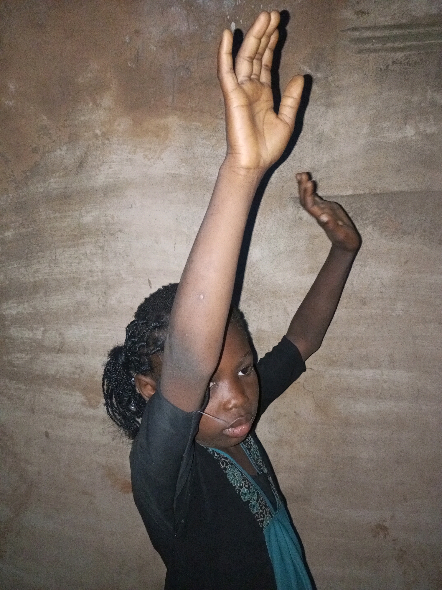 A young Black girl is pictured from the chest up, facing the camera with her arms raised. She has braided hair that is dark and pulled back. She is wearing a black shirt with a blue undershirt and an embroidered collar. The background is a textured, tan wall, which appears to be made of concrete or stucco. The lighting suggests an indoor setting, possibly at night or in a dimly lit room. Her expression is neutral. She is standing in Tempure, Nigeria.