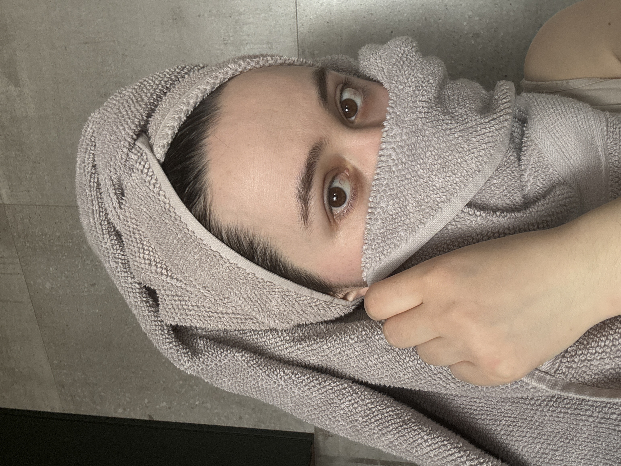A person lies on a gray tiled floor with a gray towel wrapped around their head and partially covering their face. Only their eyes are visible. The person's right hand is holding the towel. Part of a dark object is visible at the bottom left of the image. The location is not evident, but the city of Izhevsk, Russia is specified.