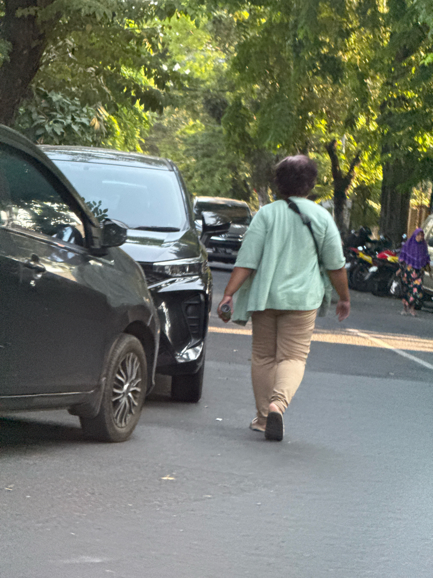 The image captures a street scene in Surabaya, Indonesia, under the warm glow of late afternoon sun. In the foreground, the left side of a dark-colored car is visible, its side profile showing a portion of the wheel and door. To its right, another black car is parked, its front end partially obscured by the first car.
Further down the street, a person is walking away from the camera, dressed in a light green, loose-fitting top and light brown, relaxed-fit pants. A dark strap crosses their right shoulder. They are carrying a cylindrical object in their left hand.
In the background, to the right of the walking person, several motorcycles are parked. Beyond the motorcycles, a woman wearing a purple hijab and a colorful floral dress is walking. The street is lined with lush green trees, their leaves illuminated by the sunlight, creating a dappled effect on the asphalt. The overall impression is a common urban scene in Surabaya, likely during a time of day when the sunlight is soft and warm.