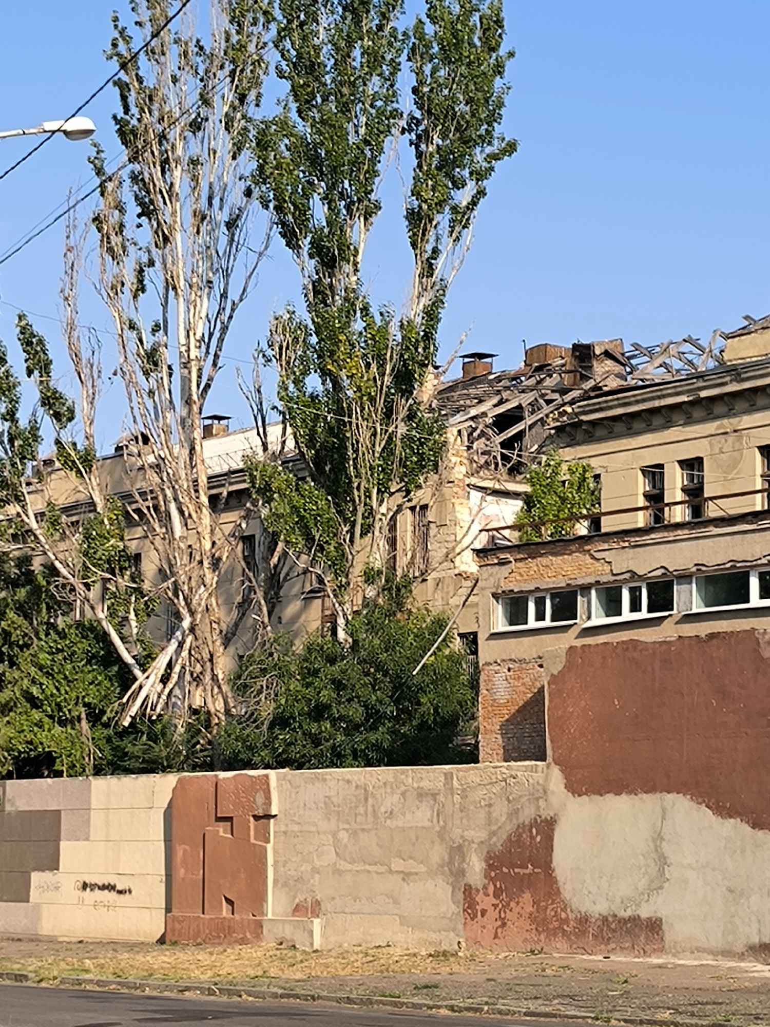 The image shows a partially destroyed building in Mykolaiv, Ukraine. The building's roof is severely damaged, with exposed beams and debris. The windows are mostly intact, but some are broken. The exterior walls are peeling and weathered, with some areas of exposed brick. In the foreground, there is a low concrete wall with some graffiti and a red, abstract sculptural element. Tall, bare trees with some green foliage stand in front of the building. The sky is clear and blue. The scene appears to be an urban environment, possibly residential or industrial. There are no people visible in the image.