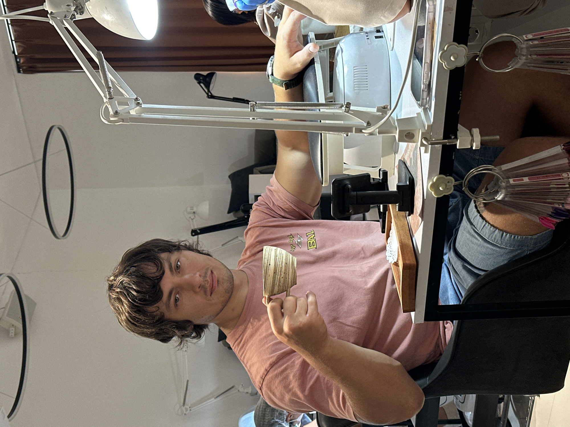 A man with brown, wavy hair holds a patterned, wooden item in his left hand. He is seated at a white table illuminated by a white, adjustable lamp. The table is cluttered with nail polish samples, a white manicure machine, and other tools. The man wears a pink t-shirt and denim shorts. The background is a bright, minimalist interior with black, circular decorative elements on the wall. The location is Kerobokan Kelod, Indonesia.