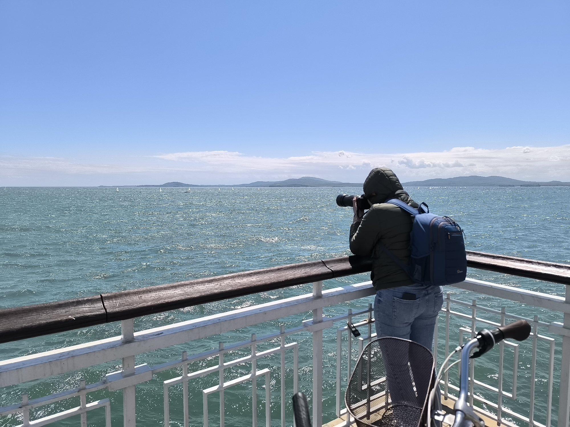 A person in a hooded jacket and jeans stands on a white railing platform with a dark wooden handrail, holding a camera with a long lens to their eye, looking out over a body of water towards some distant land masses under a blue sky. They have a blue backpack on. A bicycle is nearby on the platform. The water is turquoise and textured with small waves. The scene appears to be shot on a clear, sunny day. Small white sailboats are visible in the distance on the water. The location is likely the Burgas Pier, Bulgaria.