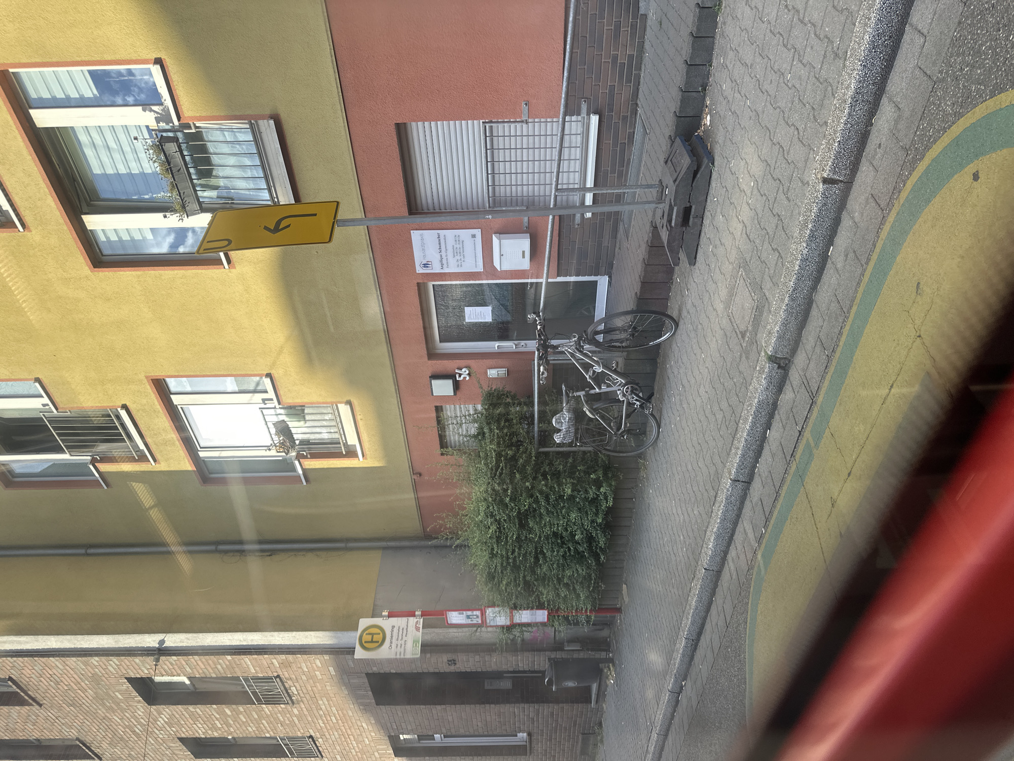 The image shows a street scene in Krefeld, Germany, likely near a residential area or small commercial district. A yellow building with pink trim dominates the right side of the frame. A street sign with a left-pointing arrow is visible. Below the sign, there is a door with the number "56" above it, and a sign for "Angelique Schumacher - Hausarztpraxis" (Angelique Schumacher - General Practice). A white mailbox is mounted to the wall next to the door. To the left of the yellow building, there is a bus stop with a sign featuring the letter "H" in a yellow circle, indicating a public transport stop. A bicycle is parked on the sidewalk in front of a dense green bush. The sidewalk is paved with interlocking stones, and a road with yellow and green markings is visible in the foreground, suggesting a crosswalk or bike lane. A brick building is visible on the far left, with windows and window bars. The lighting suggests it is daytime, with sunlight casting shadows.