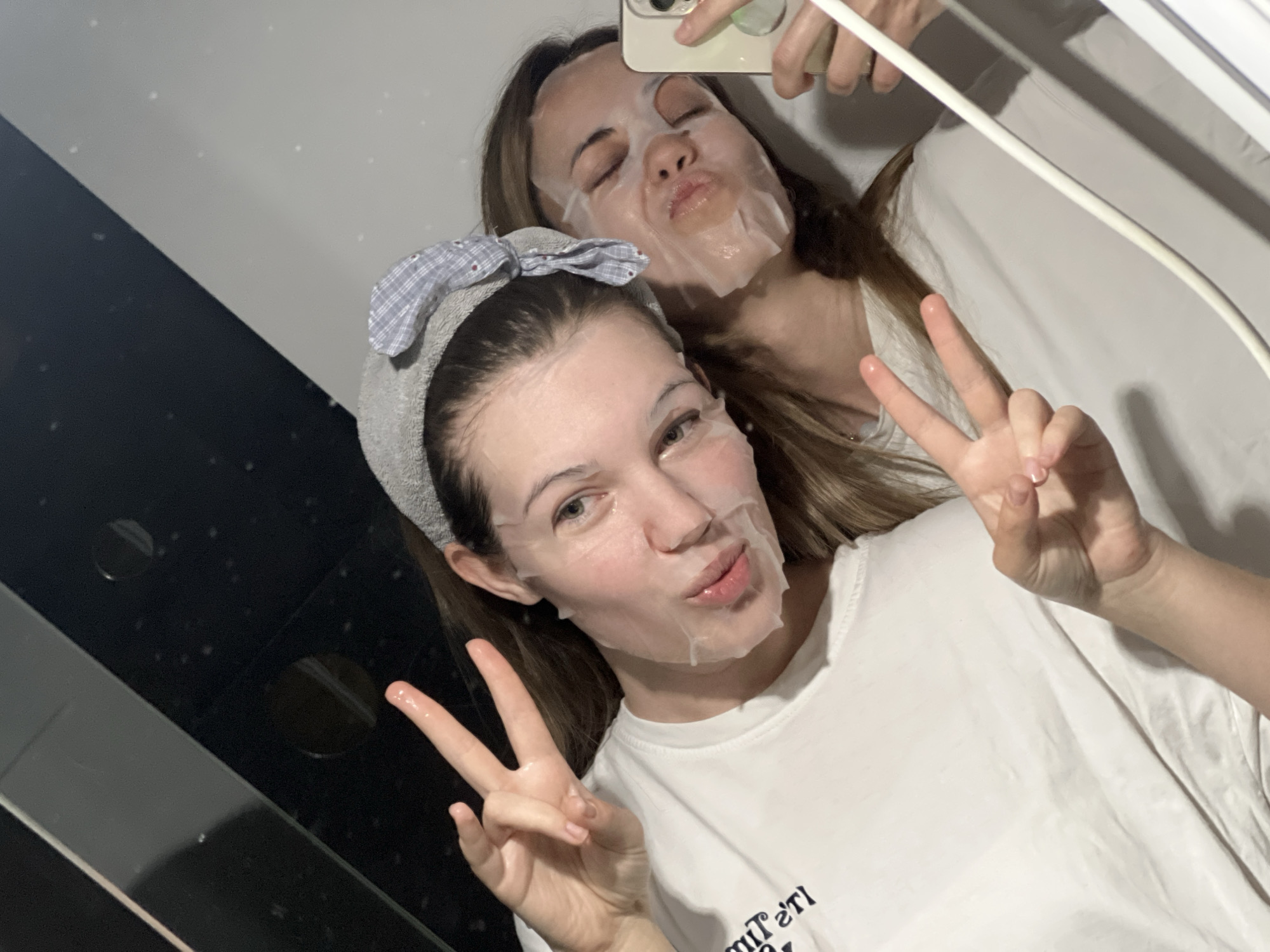 Two young women are captured in a playful mirror selfie, both wearing white sheet face masks as part of a skincare routine, likely in an indoor setting within Vozdovac Urban Municipality, Serbia.
In the foreground, one woman smiles faintly while pouting her lips and holding up both hands in a peace sign. She wears a grey terry cloth headband adorned with a light blue and white checkered bow, holding her long, light brown hair back from her face. Her white t-shirt shows partial, mirrored text that appears to be "IT'S TI" at the bottom. Her nails are neatly manicured with a light polish.
Behind her, reflected in the mirror, the second woman takes the photograph with a smartphone, visible in her hand in the upper right corner. She also wears a sheet mask and pouts her lips with her eyes closed, adding to the lighthearted mood. Her long, light brown hair cascades over her shoulders, and she is also wearing a white t-shirt.
The background of the mirror selfie reveals a diagonal division: a dark, reflective surface on the lower left, possibly a counter or part of a wall, and a lighter, off-white surface above it. A white, linear object or light fixture is visible on the far right. The overall lighting suggests an indoor setting, possibly evening or night, focused on a self-care activity. The image conveys a relaxed and friendly atmosphere.