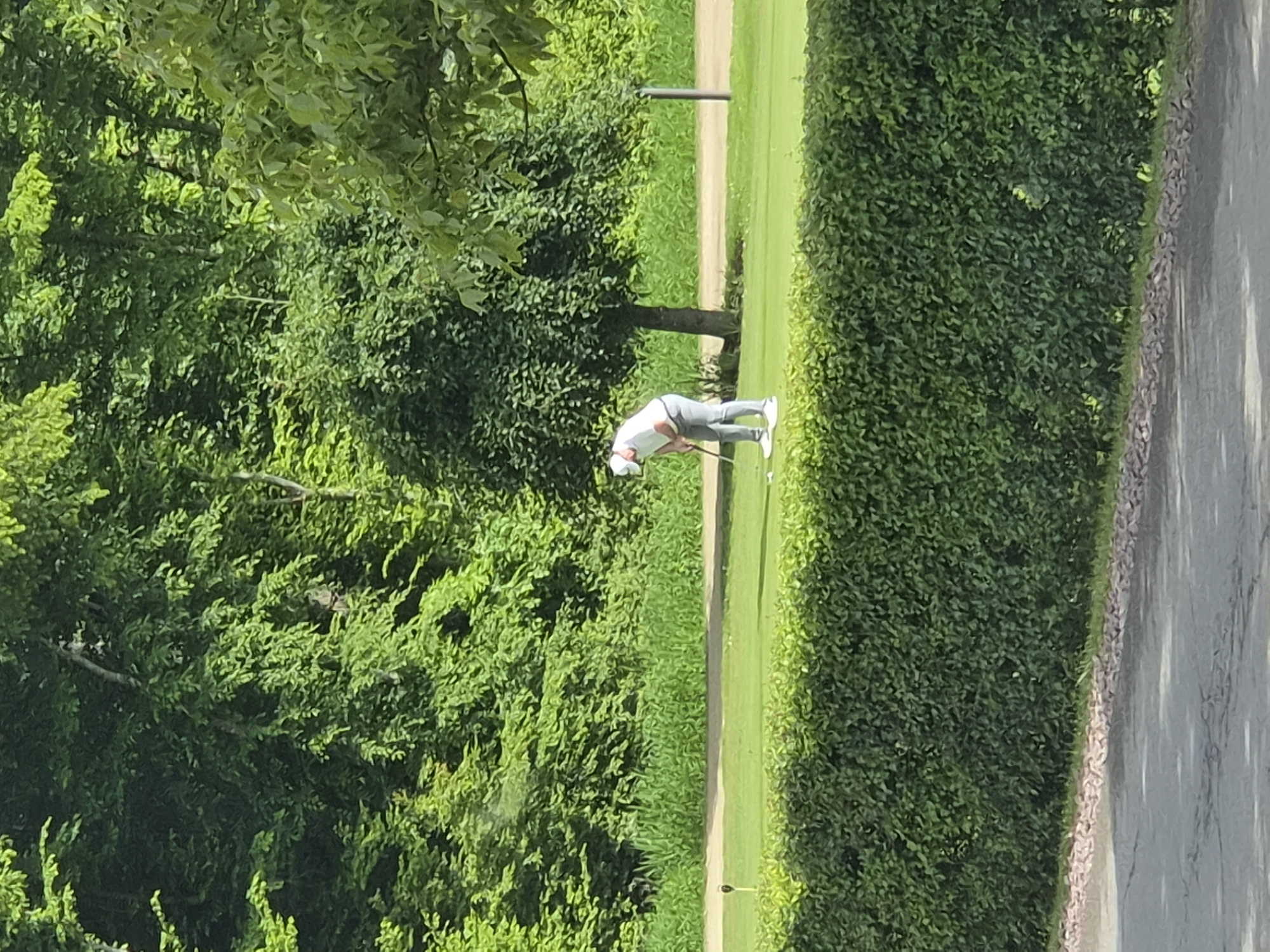 A golfer dressed in white hat, white shirt and grey pants, is playing golf on a green golf course in Lenningen, Luxembourg. He is swinging a golf club. A tall green hedge lines the right side of the golf course, while a dense green forest is on the left. A paved area is visible along the right side of the image.