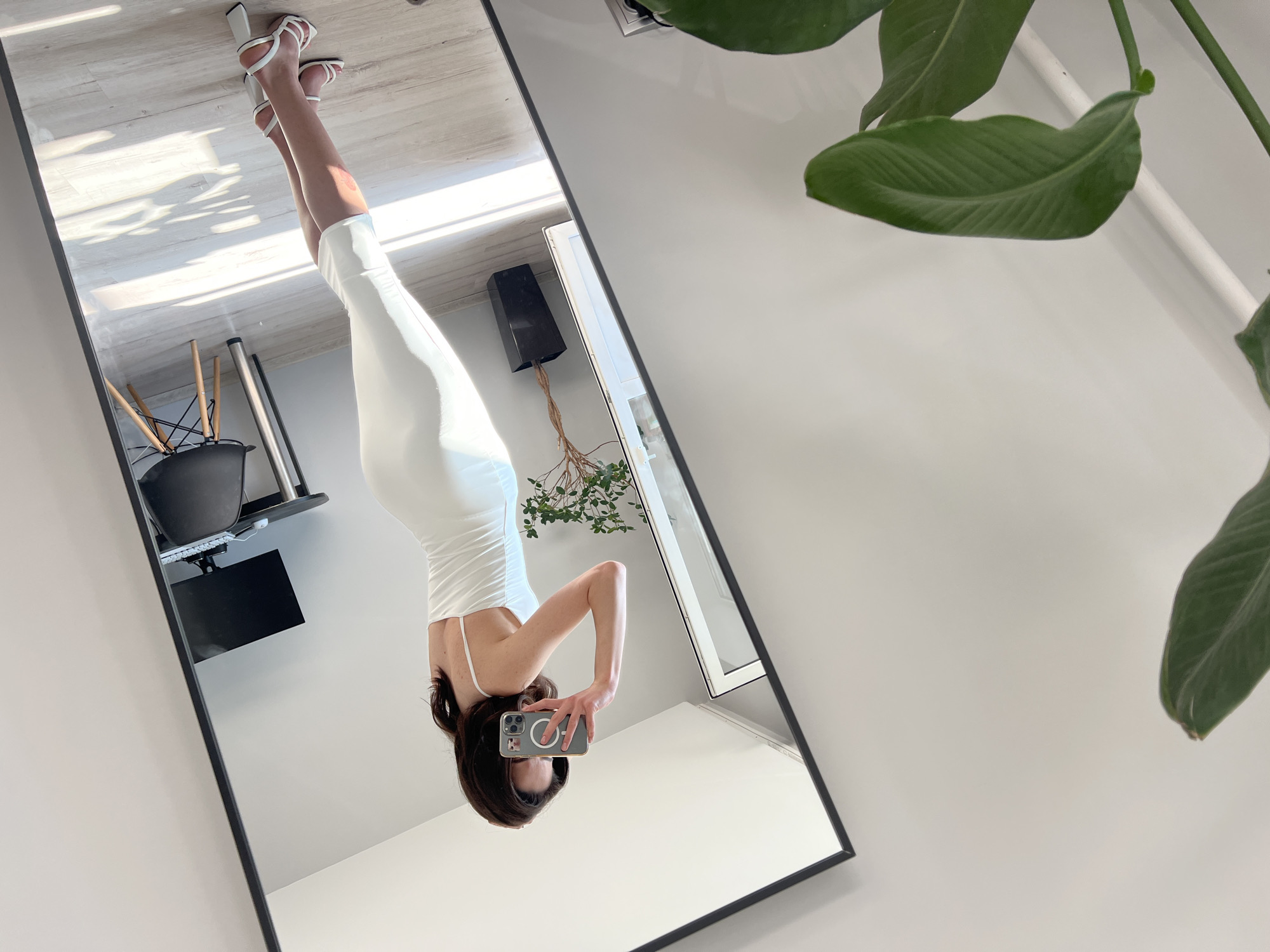 A woman in a white slip dress and white heeled sandals is taking a mirror selfie in a room. She is holding a smartphone in her right hand and has her left hand touching her hair. The room appears to be a modern, minimalist space with white walls and light-colored flooring. A large mirror is prominent in the scene, reflecting the woman and parts of the room. In the reflection, a desk with a computer monitor and a black chair are visible. A potted plant with green leaves is situated to the left of the woman, and another similar plant is visible in the mirror's reflection, placed in a black planter. The lighting suggests daylight, with sunlight casting shadows on the floor. The scene does not contain any identifiable landmarks or elements that would place it specifically in Ternopil, Ukraine.