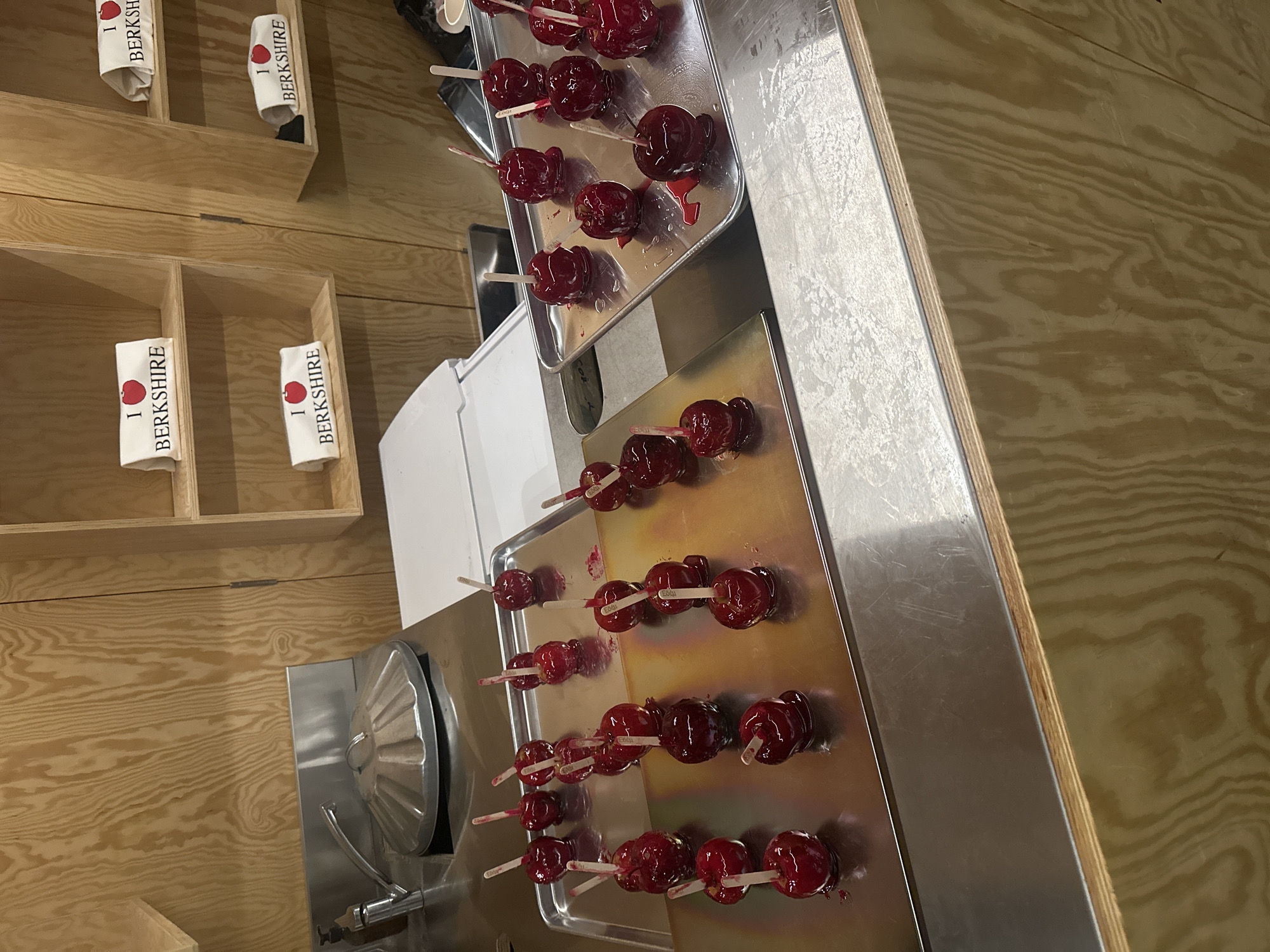 This image captures a display of numerous red candy apples set against a wooden and metallic background, likely in an indoor commercial setting such as a market or fair stall.
The primary subjects are rows of shiny, crimson-red candy apples, each impaled on a wooden stick. These are meticulously arranged on two reflective, rectangular metal trays, which appear to be made of stainless steel. Some apples show small drips of the candy coating on the trays, indicating their fresh preparation.
In the upper left portion of the image, several white rectangular cloths or napkins are folded and placed within light-colored wooden shelves. Each cloth clearly displays the text "I ❤️ BERKSHIRE," with the heart symbol represented by a red apple graphic. There are at least four such cloths visible across two shelves. The wooden shelving itself is simple, with multiple empty compartments.
The overall setting features a prominent light-toned wooden wall or counter, showing a distinct wood grain, which serves as the backdrop. A sturdy metal surface, possibly a counter or part of a food preparation unit, runs along the right side and bottom of the image, supporting the candy apple displays. Part of what appears to be a large, metallic, ribbed lid (possibly from a machine or container) is visible on the lower left.
Given the text "I ❤️ BERKSHIRE" and the provided location context of London, United Kingdom, this scene likely takes place at an event or establishment celebrating or selling goods related to the Berkshire region, which is a county neighboring London. The image depicts a moment of display, possibly before or during an event, showcasing these confectionery treats and regional merchandise. The lighting is even, suggesting an indoor environment without direct harsh sunlight.