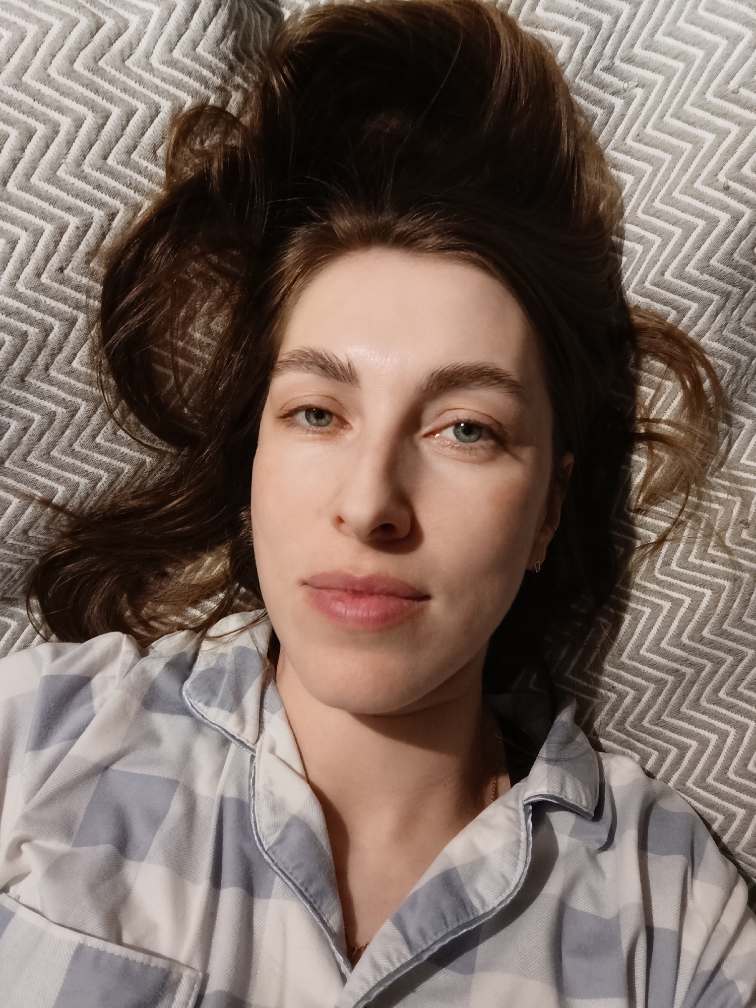 A woman with brown hair and light eyes is lying on a patterned grey and white blanket or bedspread, wearing a blue and white checkered pajama top. Her hair is spread out around her head, and she is looking directly at the camera. The location is not discernible from the image, and there is no evidence to suggest the city of Chaikovsky, Russia.