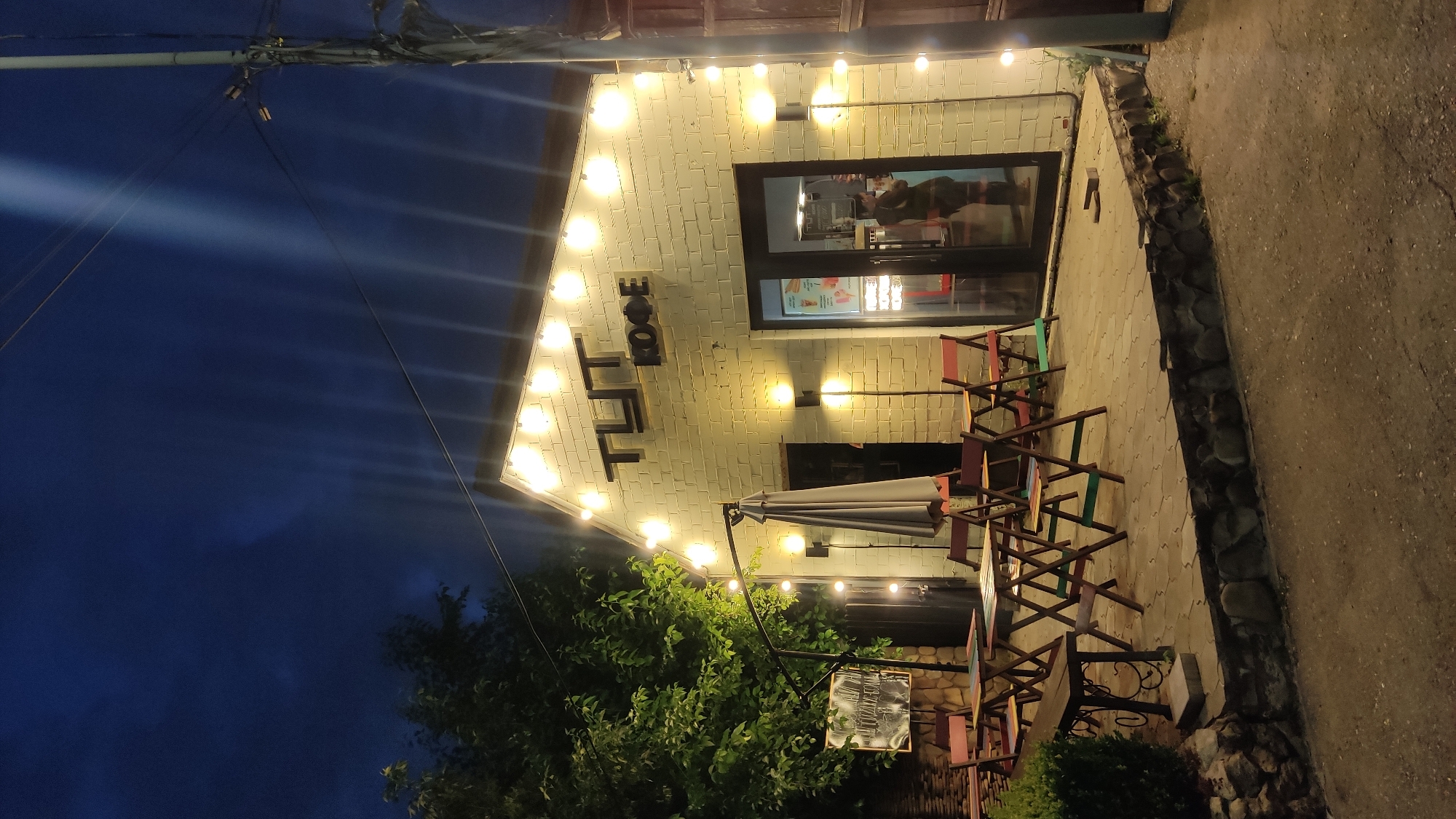 This is a nighttime exterior shot of "Tut Kofe," a cafe located in Krasnodar, Russia. The building is made of white brick and is illuminated by string lights along the roofline and spotlights on the facade. A sign above the entrance reads "TUT KOFE" in black lettering.
In front of the cafe is an outdoor seating area with several small tables and chairs arranged on a paved patio. A large gray umbrella stands folded near the entrance. A person is visible inside the cafe through the glass doors. To the left of the building, there is a blackboard sign and lush green trees. The ground in front of the patio is a gravel or dirt path. The sky above is dark blue, indicating it is nighttime.