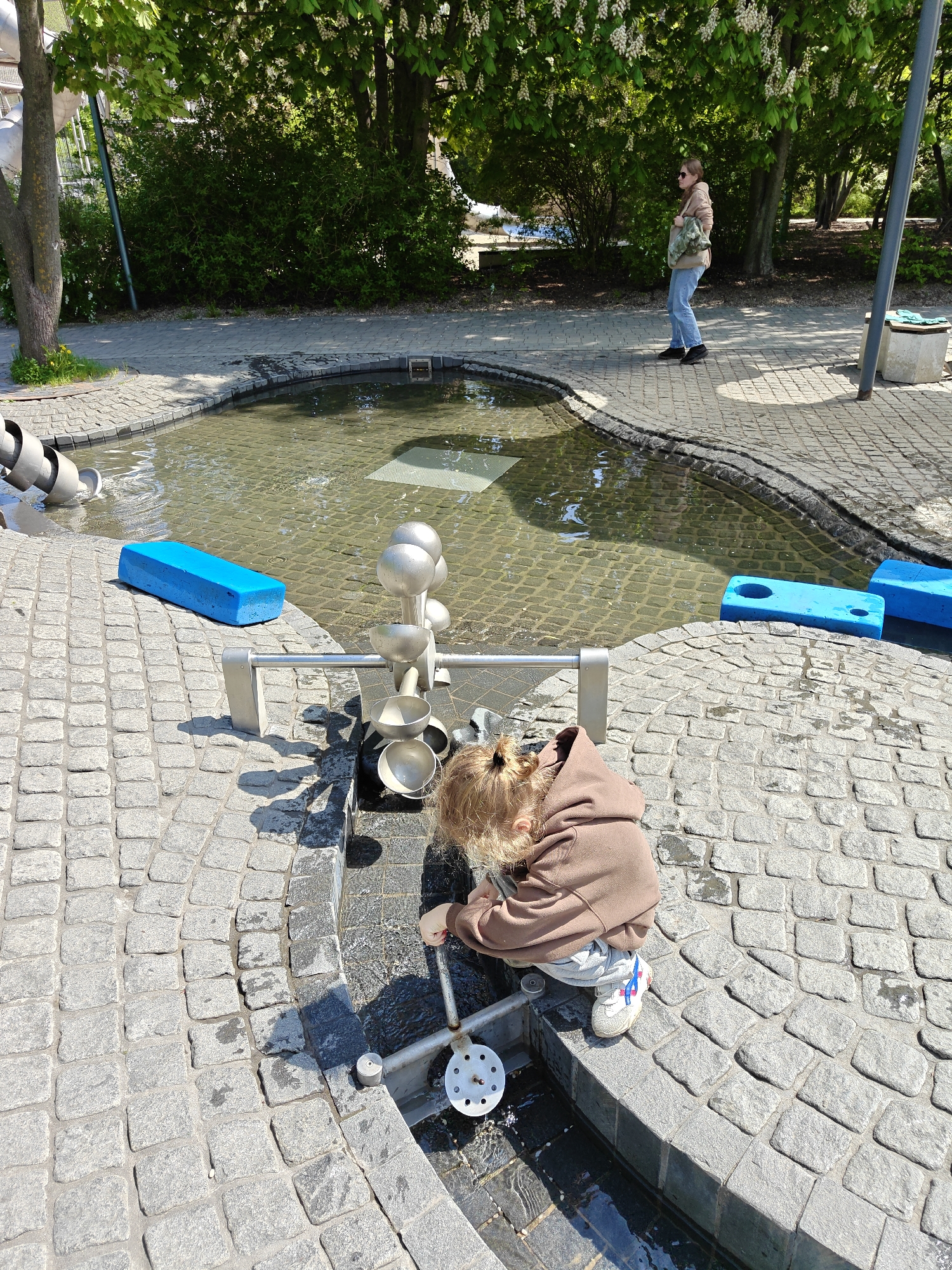 A wide shot shows a young child playing at a water feature in what may be Gorky Park in Moscow, Russia. The water feature is a shallow pool surrounded by gray cobblestones, with various interactive elements, including a series of interconnected metal bowls and a hand-operated pump that the child is using. Several bright blue foam blocks are positioned around the pool.
The child is kneeling near the water, wearing a brown hooded sweatshirt, gray pants, and sneakers. They are focused on the pump mechanism, presumably trying to make the water flow.
In the background, a woman in jeans and a tan jacket walks on a cobblestone path. Trees and bushes provide a green backdrop, and the sky appears sunny and bright. A light post stands near the walking path in the background.