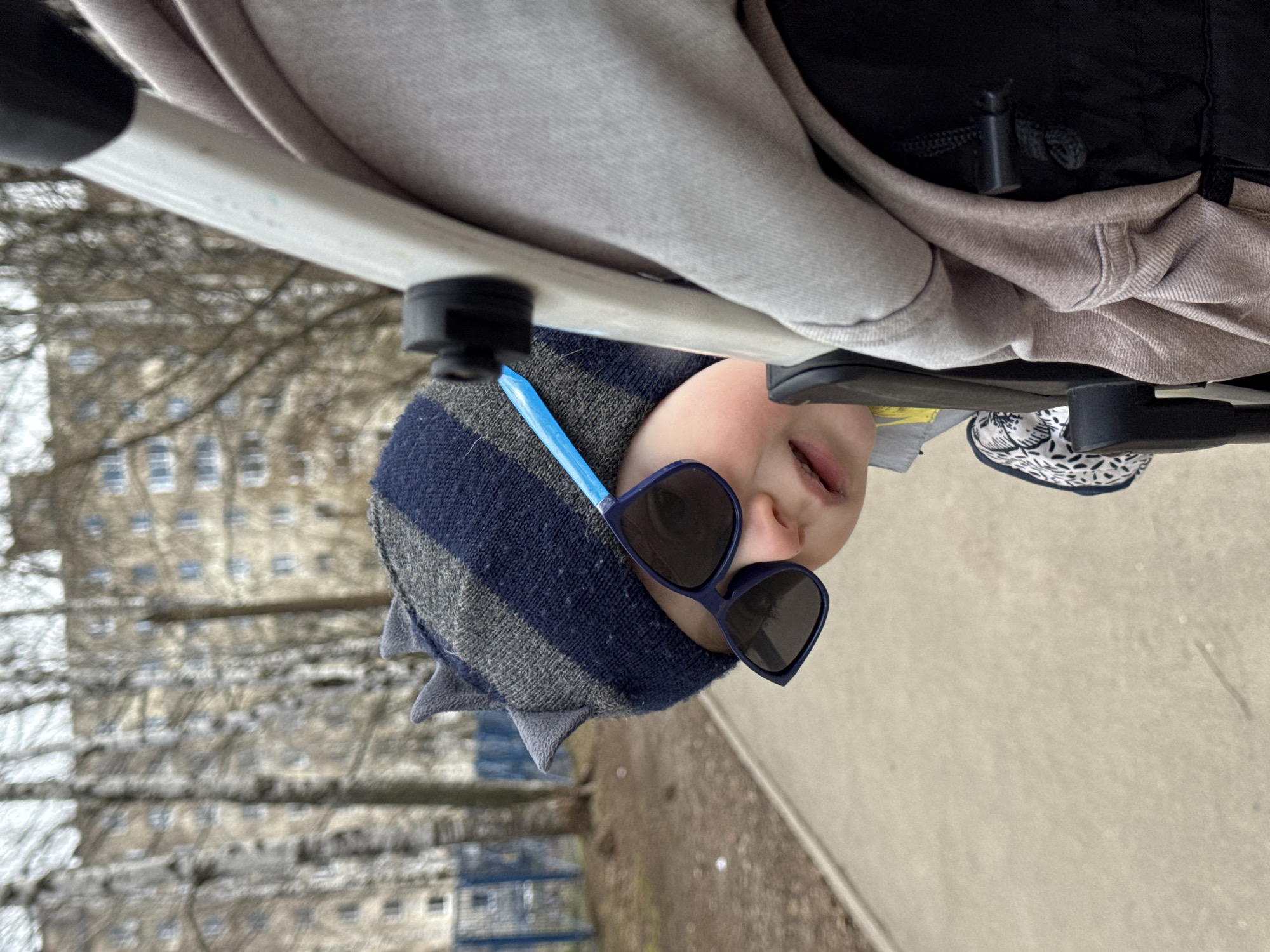 A young child is captured in an outdoor setting, positioned in a stroller or pram and looking upwards towards the camera. The child wears a distinctive blue and grey striped hat adorned with small, pointed features, resembling spikes or ears. Large, dark sunglasses with blue frames cover the child's eyes, adding a playful element. A light blue straw or similar thin object is visible near the child's head, seemingly tucked into the hat or sunglasses. The child's mouth is slightly ajar.
The upper portion of the image features the light-colored frame and grey fabric of the stroller. In the background, numerous bare trees stand, indicating a cooler season such as late autumn, winter, or early spring. Behind the trees, several multi-story buildings with visible windows can be discerned, consistent with an urban environment like Izhevsk, Russia. The ground appears to be a light-colored paved path or sidewalk. The lighting suggests it is daytime, possibly under an overcast sky. There is no visible text in the image.