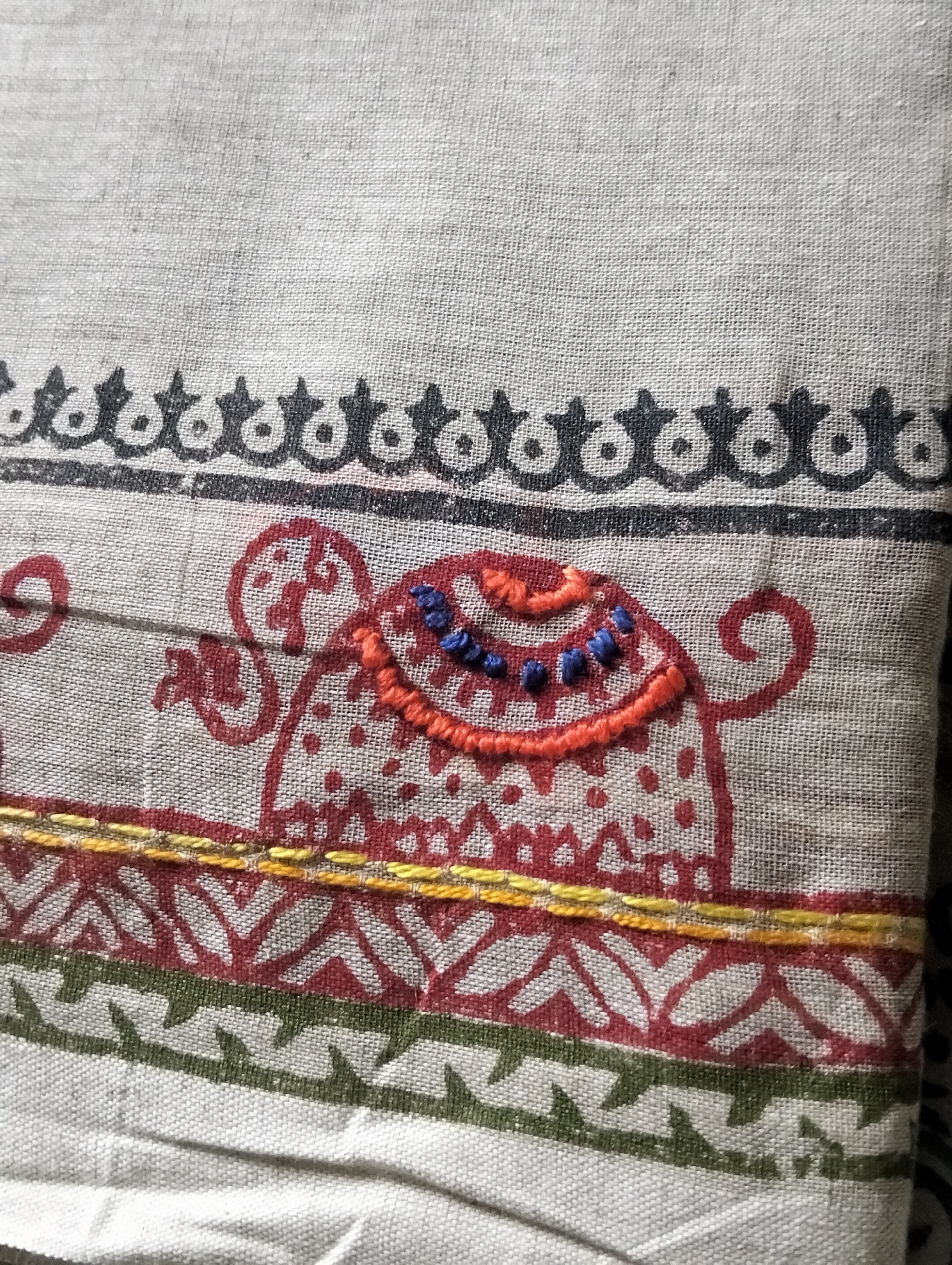 The image shows a close-up of a piece of embroidered fabric with traditional patterns and motifs. There is a black ornamental border at the top. Below, there is a red stylized animal figure with an orange, red, and blue embroidered saddle. A yellow line borders the base of the animal. Below that, there is a red and white zigzag pattern and a green zigzag pattern. The fabric has a beige or light gray background. It is not possible to determine the location as Dhaka, Bangladesh from the image.