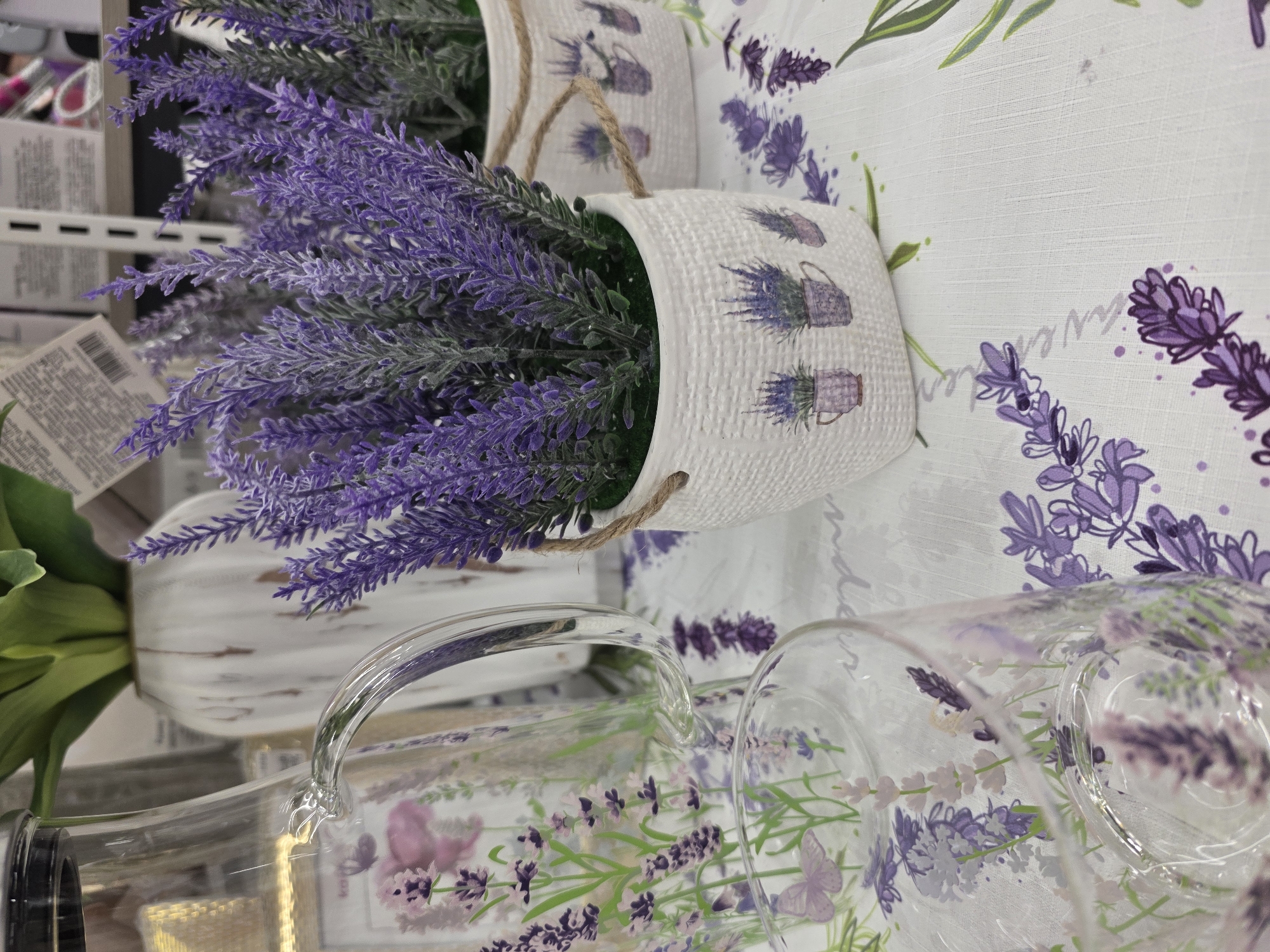 The image displays a retail presentation of lavender-themed home goods within a store in the city of Chaikovsky, Russia. In the left foreground, a clear glass pitcher with a black lid and a curved transparent handle is visible. Its surface is decorated with purple lavender sprigs, green leaves, and light purple butterfly illustrations. Below and to the right of the pitcher, the rim of a clear glass tumbler, similarly adorned with lavender and butterfly motifs, is partially visible. Centered in the mid-ground are two artificial lavender plants, each contained within a white, basket-weave textured pot featuring small illustrations of lavender plants in cups and handles made of twine. The artificial lavender consists of numerous purple-flowered sprigs with green stems. All these items are arranged on a white tablecloth patterned with repeating designs of purple lavender sprigs, green leaves, and the word "lavender" in script. The background is out of focus, revealing white metal retail shelving stocked with various products and white price tags, along with a white, distressed-style vase holding a green artificial plant. No individuals are present in the scene.