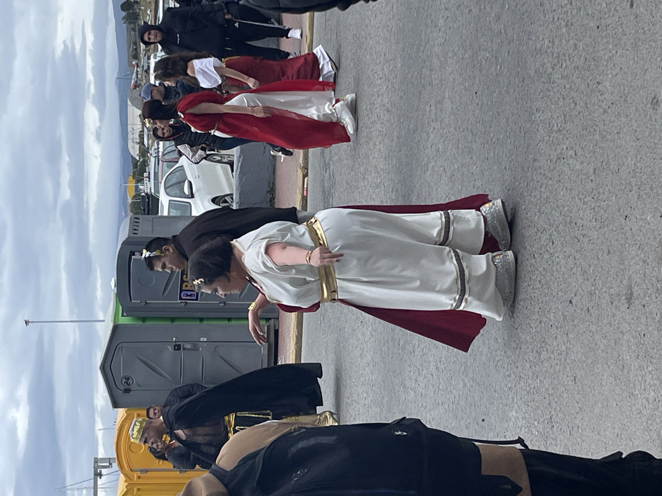 The image captures an outdoor event featuring a group of people dressed in ancient Roman or Greek-inspired costumes, likely a parade, festival, or performance. The scene is set on asphalt pavement, with several individuals lined up, and portable toilets visible in the background, suggesting a public, temporary event. The sky is overcast, indicating a cloudy day.
In the immediate foreground, a person is partially visible wearing a black garment with what appears to be a brown or gold sash. To their right, a woman is seen in a flowing white gown with a red sash or cape draped over her shoulders. She wears a gold belt and a gold wreath on her head. Her shoes are notably sparkly silver. She is bent slightly at the waist, with her arms raised slightly and her fingers curled, suggesting a dance move or part of a performance.
Behind this woman, slightly to the left, is another man in a black long-sleeved shirt, also wearing a gold wreath on his head. Further back and to his right, two gray-green portable toilets are positioned. To the left of these toilets, a yellow portable toilet is partially visible.
Continuing further into the background, a line of individuals dressed in similar ancient costumes is visible. The closest ones are wearing red and white gowns with red capes, some also adorned with gold elements and gold wreaths. One woman in a red and white costume is looking towards the left, while others are oriented towards the front. In the mid-ground, a white car is parked behind the costumed participants.
The farthest background shows more people, some in modern casual attire (black jackets, sweatpants, sneakers), standing and observing the event. Beyond them, an urban or semi-urban landscape is visible with buildings and what appear to be distant mountains under the cloudy sky. The overall impression is one of a cultural celebration or historical reenactment taking place in an open, public area. No discernible text is clearly visible on any objects or clothing in the image.