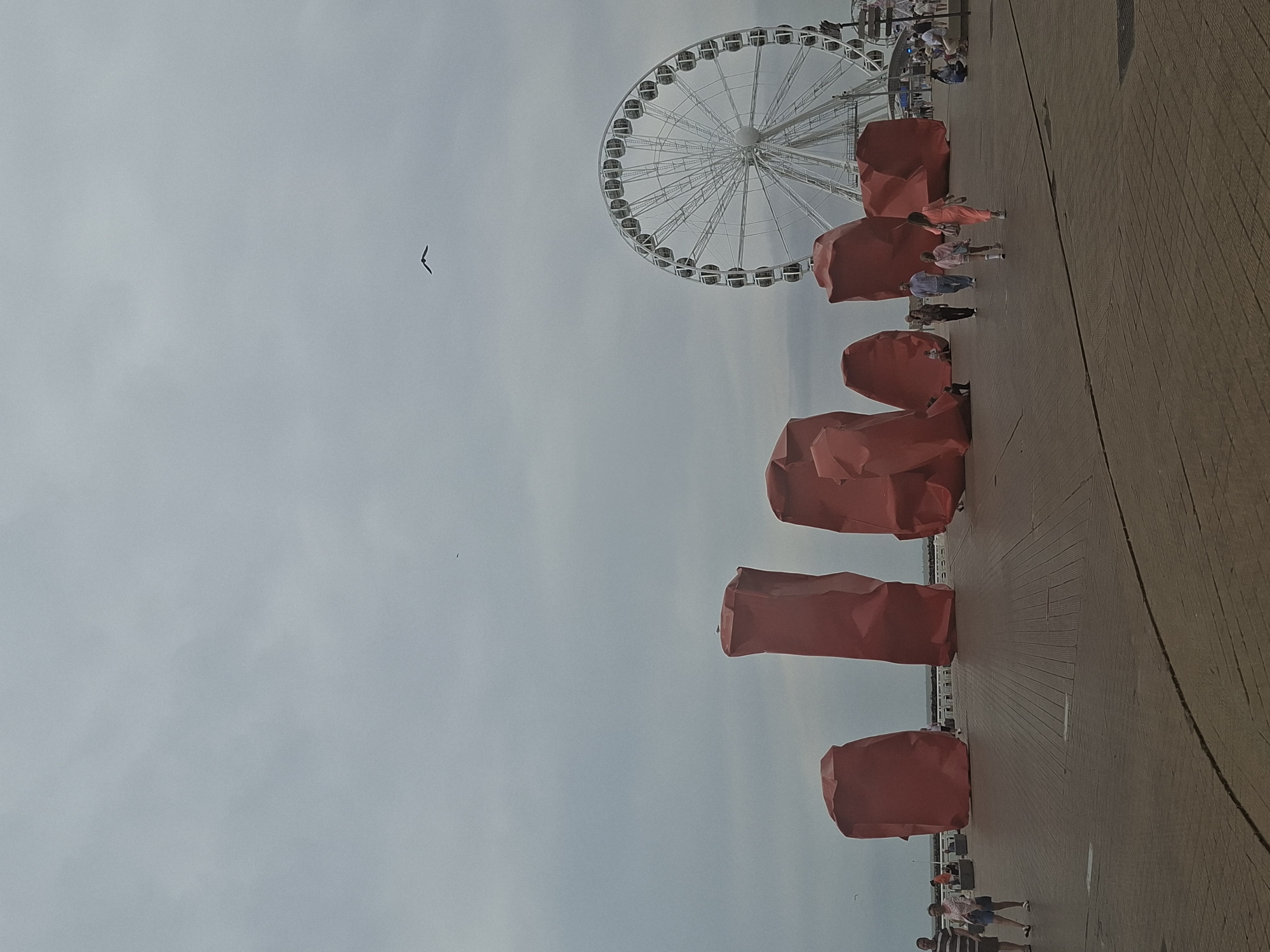 The image depicts the waterfront promenade in Ostend, Belgium. Dominating the foreground are several large, bright red abstract sculptures, some resembling crumpled forms. Beyond these sculptures, a white Ferris wheel stands prominently against a cloudy sky, suggesting an amusement area. Several people are visible on the promenade, walking and socializing. A bird is captured in flight in the upper portion of the frame. The ground is paved with wooden planks.