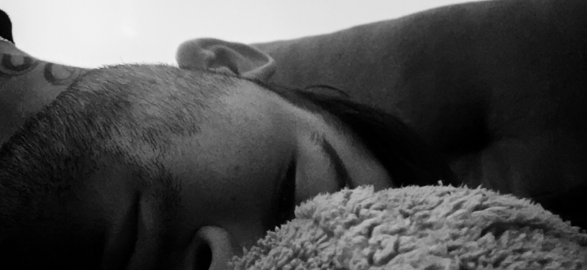 A black and white, close-up shot depicts a person sleeping, resting their face on a soft, textured surface. The person's face is turned towards the viewer, with closed eyes and visible ear. The texture of their hair is visible and appears short. A pattern, possibly a tattoo, is noticeable on the back of the head near the hairline. The background is out of focus but suggests a blanket or other soft surface the person is lying on. There is no visual information to suggest this photo was taken in Plovdiv, Bulgaria. The scene is indoors.