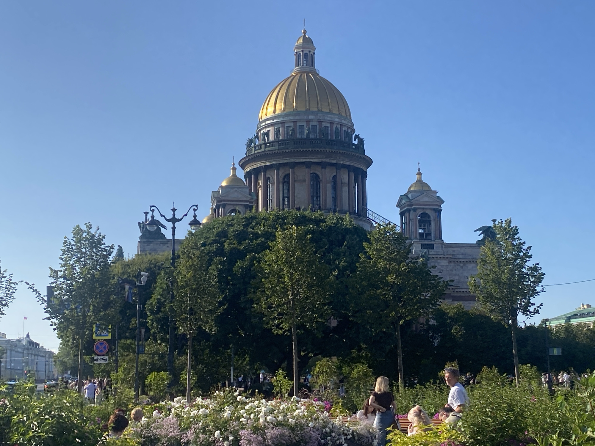 This image depicts Saint Isaac's Cathedral, a prominent landmark in Saint Petersburg, Russia, on a clear, sunny day. The massive neoclassical cathedral, with its iconic golden dome, dominates the background. In the foreground, a lush green park area is filled with a variety of flowering plants, including white and pink flowers, and dense foliage. Several people are visible in the park, some sitting on benches, others walking or standing. A man in a white t-shirt is seated on a bench, looking towards the camera, while a woman with blonde hair in a dark top is standing near him. Other individuals are partially visible in the surrounding greenery. The scene is framed by trees, and streetlights are present along a pathway. In the distance, other buildings of Saint Petersburg can be seen under the clear blue sky.