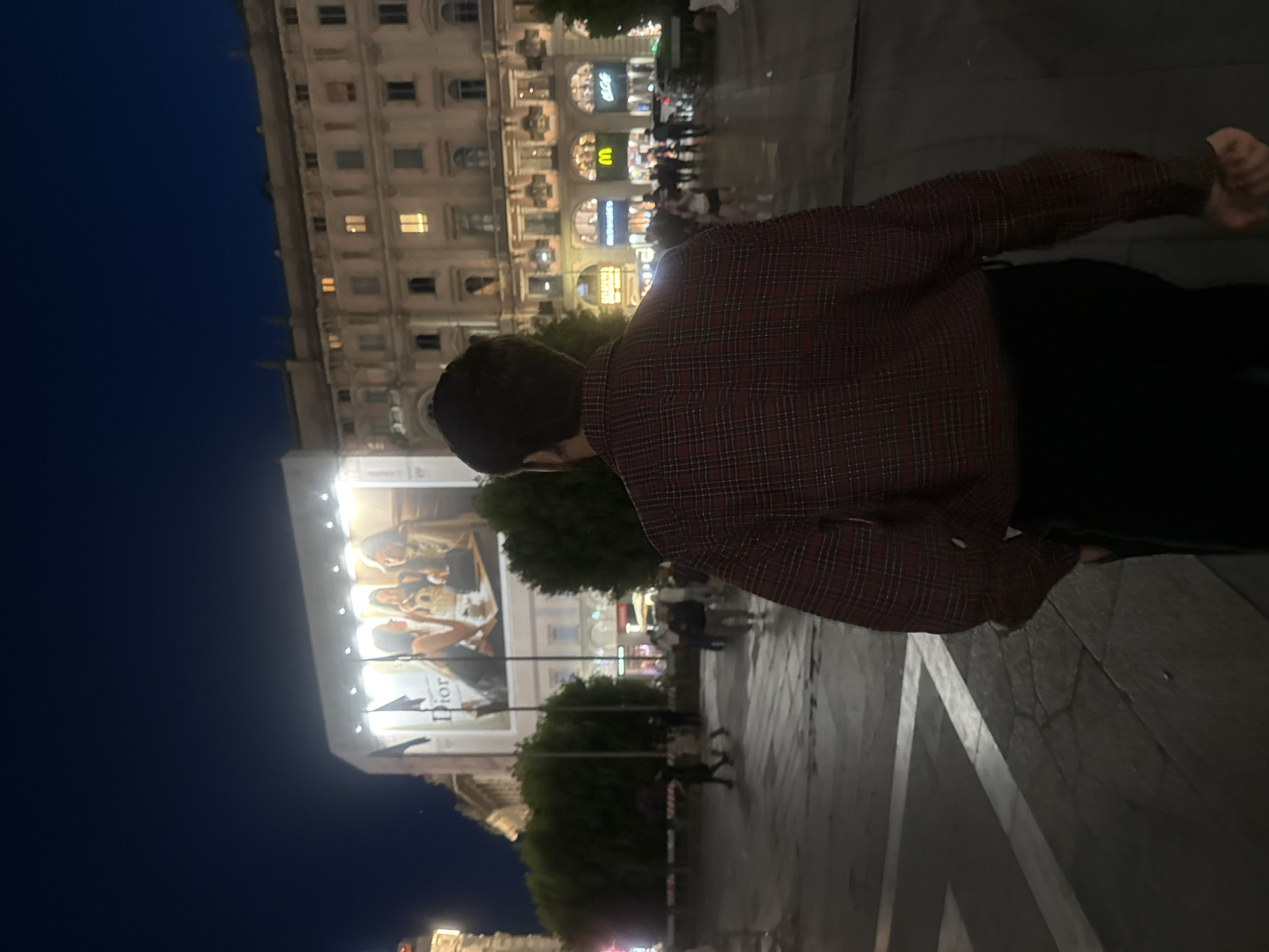 At night, a person in a checkered shirt stands in Milan's Piazza del Duomo. The Duomo is in the background, along with storefronts and people gathered in the square. A large Dior advertisement is visible. The ground appears wet, reflecting ambient light.