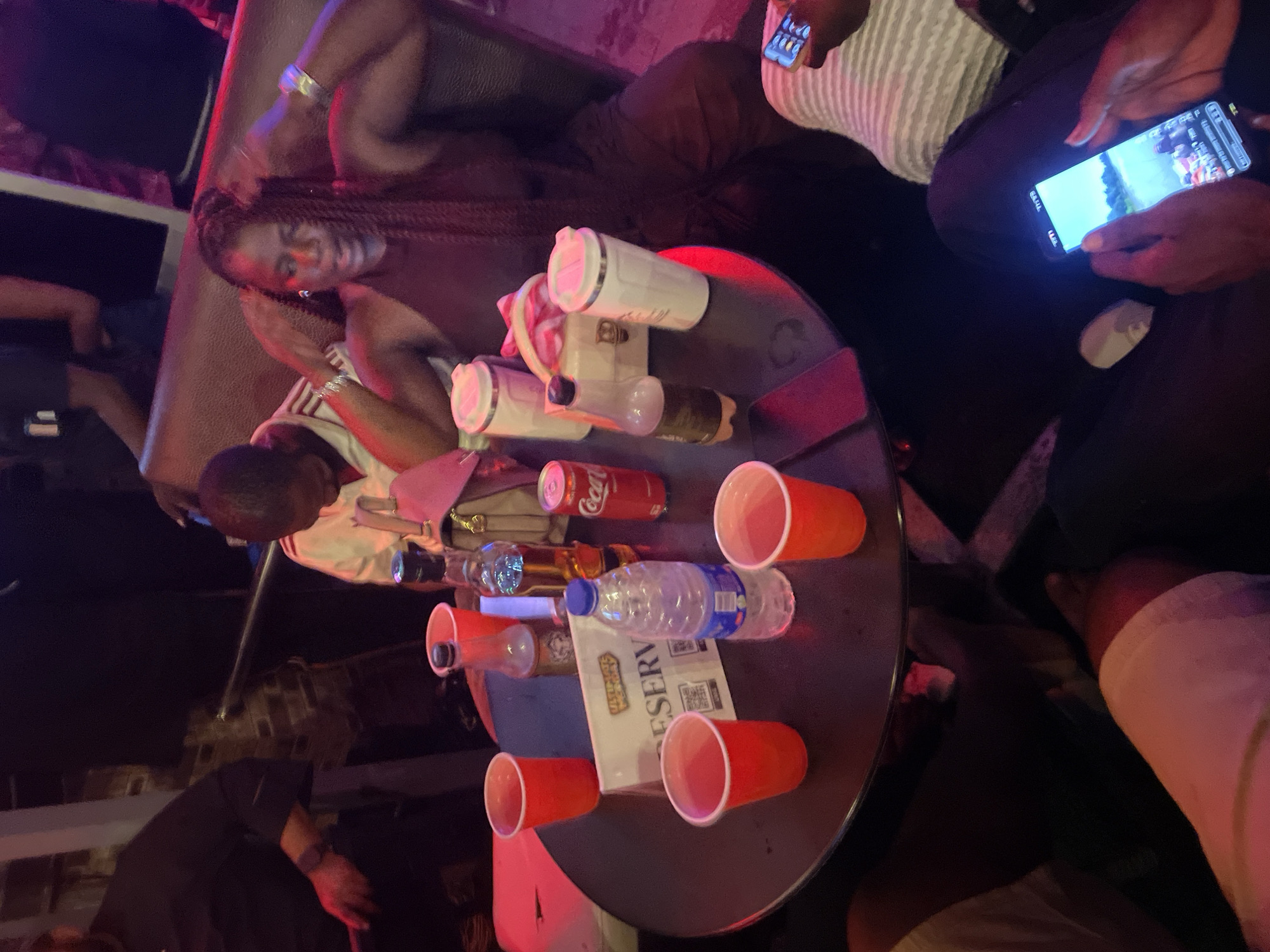 The image depicts a group of individuals seated around a circular table in a dimly lit establishment, likely a lounge or club, located in Guzape, Nigeria. The ambiance suggests a social gathering, possibly a night out. A woman with braided hair is prominently featured in the foreground, her hands raised near her head. On the table, an array of items includes multiple red disposable cups, a can of Coca-Cola, a bottle of water, a bottle of alcohol, and two white tumblers with lids. A "RESERVED" sign is placed on the table, indicating a designated area. Other individuals are partially visible, some seated and some standing in the background, though their faces are not clearly discernible. One person in the lower right corner is holding a smartphone displaying a video or image. The overall setting appears casual, with patrons engaged in conversation or enjoying their drinks.