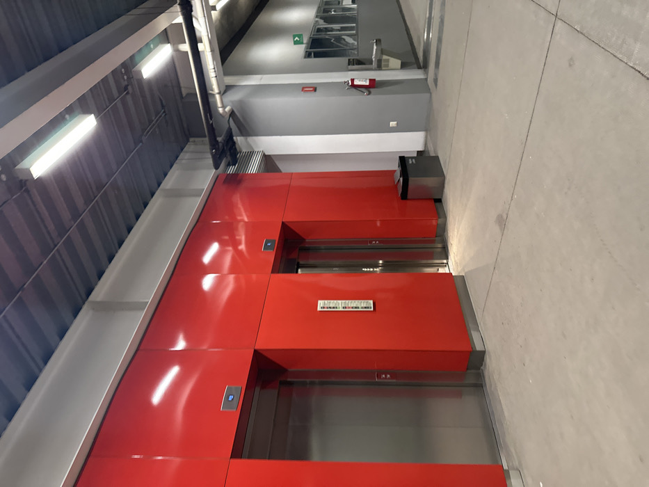 The image shows a row of red elevator doors in a modern building. Three elevator openings are visible, each framed by vibrant red panels. The doors themselves are a metallic silver color. To the left of the elevators is a metallic gray wall with overhead lighting. To the right, there is a gray concrete floor. The perspective is slightly skewed, with the image appearing to be taken from an elevated angle.