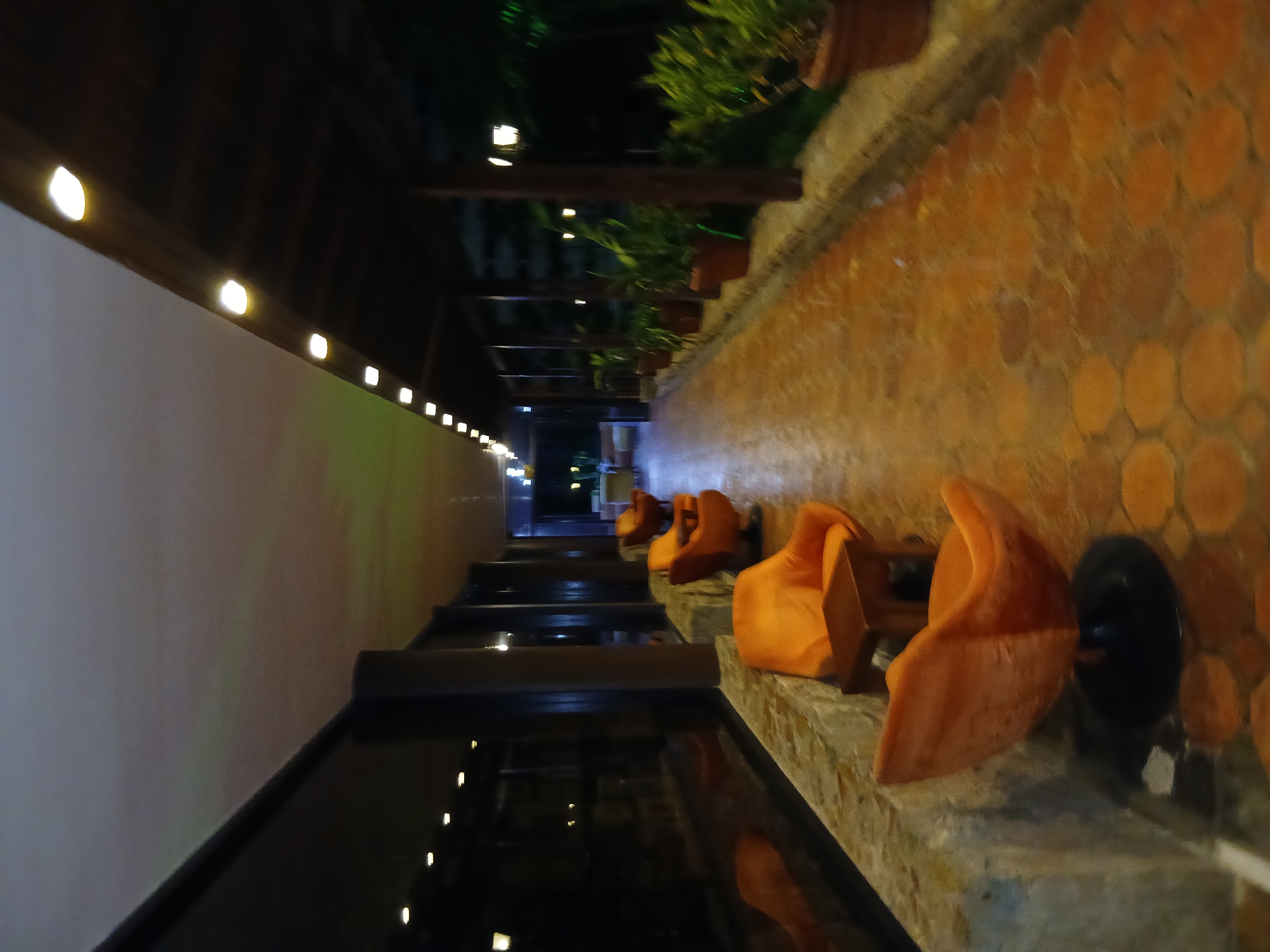 The image depicts an outdoor or semi-outdoor pathway at night, likely within a resort, hotel, or restaurant setting in Playa El Agua, Venezuela. The scene is illuminated by numerous artificial lights, creating a warm and inviting atmosphere.
Along the upper left side of the frame, a dark wooden or structural beam features a series of small, bright, warm-toned lights embedded within it, casting light downwards. Below this, a light-colored wall extends, with reflections of distant lights visible on a dark, reflective surface, possibly a body of water or a highly polished floor, running parallel to the wall.
On the right side of the pathway, several distinct, curved orange seats or chairs are arranged along a low stone or concrete ledge. These seats appear modern and comfortable. The ground immediately adjacent to these seats is covered with round, terracotta-colored tiles, forming a textured pattern. Further to the right and receding into the background, potted plants with green foliage are visible, suggesting a garden or landscaped area. A dark, possibly round object, resembling a speaker or umbrella base, is positioned near one of the orange seats.
The absence of people suggests a quiet moment, emphasizing the serene and peaceful ambiance created by the warm lighting and comfortable setup. The time of day is clearly night, indicated by the sole reliance on artificial illumination. There is no visible text in the image.