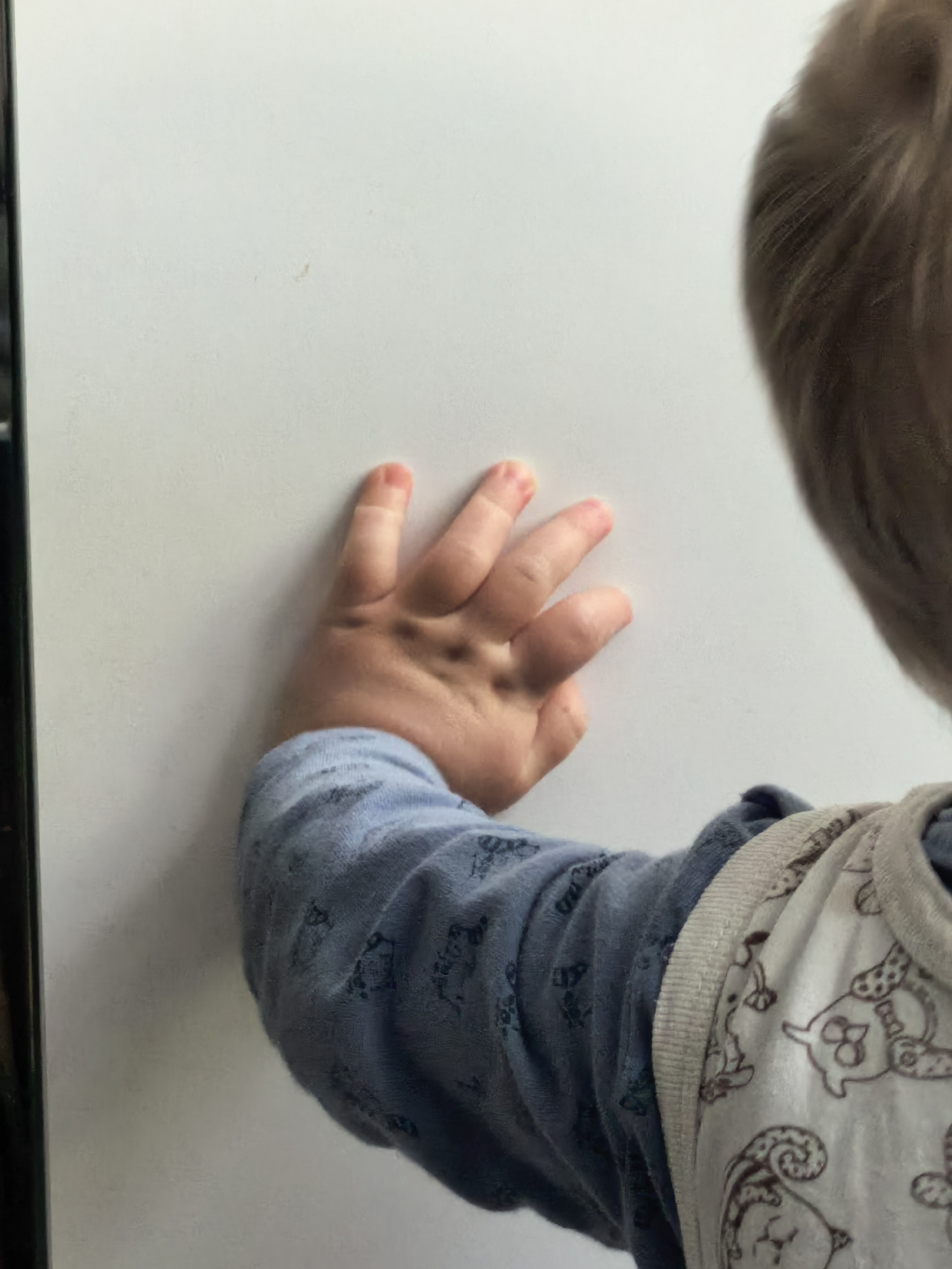 An overhead, slightly angled shot shows a young child from the back, primarily focusing on their left arm and hand against a plain white background. The child's light-colored hair is visible on the right side of the frame. They are wearing a long-sleeved light blue garment with a subtle dark pattern, possibly depicting animals or abstract shapes. Over this, a light-colored, possibly beige or off-white, sleeveless top or bib is worn, featuring a pattern of darker, cartoonish animals, which appear to be owls or similar creatures.
The child's left hand is pressed against the flat white surface, with fingers splayed out. The skin of the hand appears soft and youthful, typical of a baby or toddler. A dark vertical line runs along the left edge of the image, suggesting an adjacent object like a door frame or the edge of an appliance. The overall setting appears to be indoors, likely in a home in Sterlitamak, Russia. There is no visible text in the image.