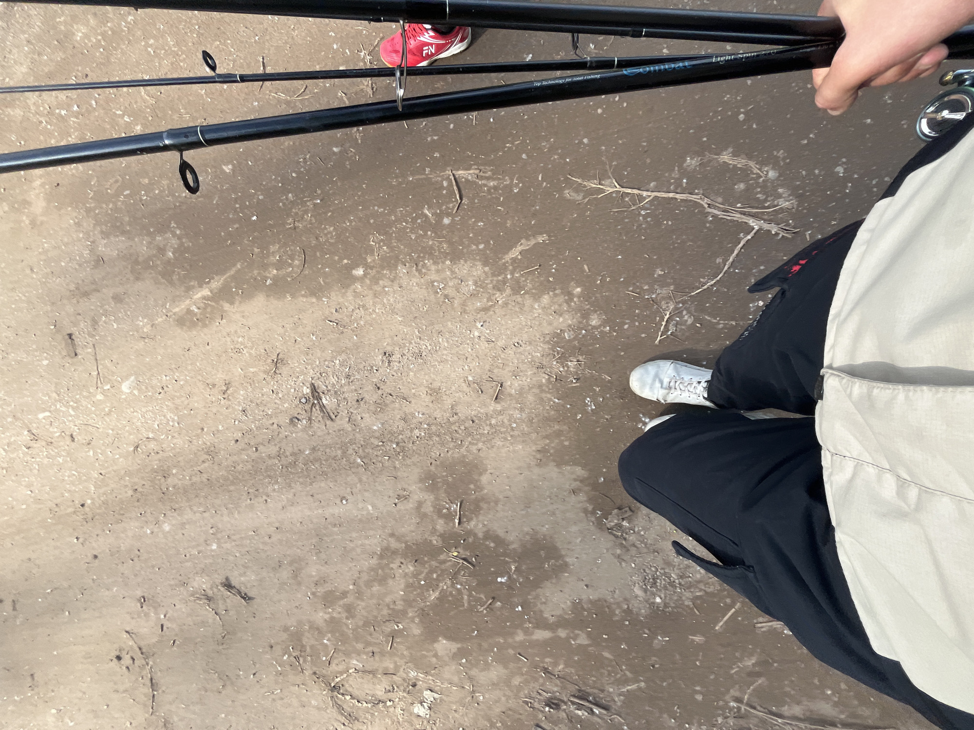 The image shows a person standing on a mud-covered ground surface. The person is wearing dark pants, a light-colored shirt, and white shoes. They are holding two black fishing rods in their hand. One rod has the word "Combat" written on it. Another person's red sneaker with the letters "FN" can be seen in the background near the fishing rods. The surrounding environment appears to be a muddy ground surface with scattered twigs and debris. The location is Chaikovsky, Russia.