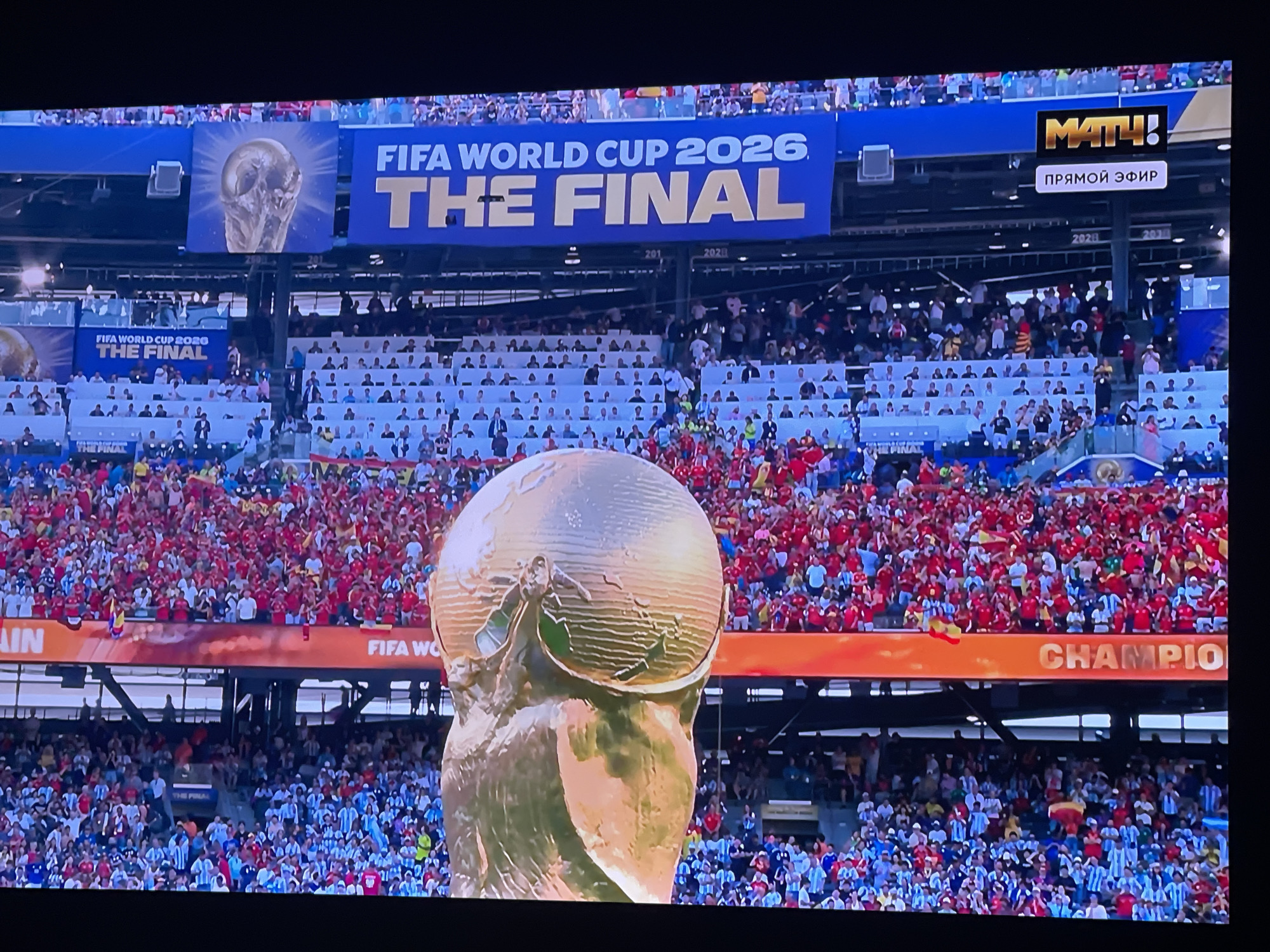 The image displays a stadium crowd watching a large screen broadcasting the FIFA World Cup 2026 Final. The screen prominently features the FIFA World Cup trophy and the text "FIFA WORLD CUP 2026 THE FINAL." A Russian broadcasting logo "МАТЧ!" and the text "ПРЯМОЙ ЭФИР" (Live Broadcast) are also visible on the screen. The crowd is densely packed, with many individuals wearing red and white or blue and white attire, likely supporting different national teams. The stadium seating areas are visible in the background, filled with spectators. The overall scene suggests a major sporting event being broadcast live to a large audience. No specific location within the city of Stary Oskol, Russia is identifiable from the image.