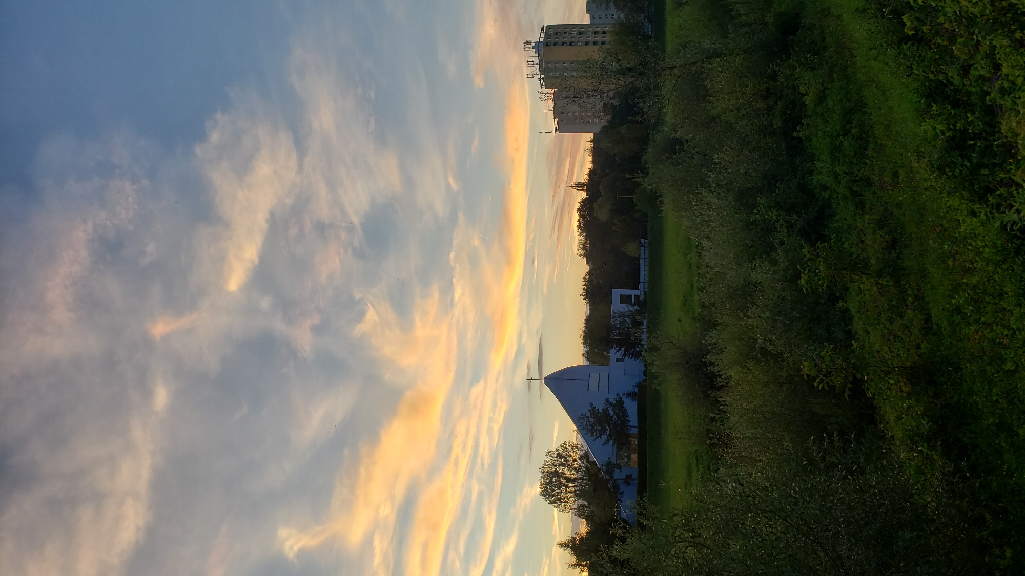 An outdoor, vertical, eye-level shot captures a suburban landscape at sunset. In the mid-ground, a modern, white church with a distinctive triangular roof and a cross on its peak stands to the left, surrounded by trees. Adjacent to the church, to the right, is a white building with rectangular windows and a dark, flat roof. Further to the right in the mid-ground, two apartment buildings rise: a lighter colored one on the left with multiple floors and many windows, and a taller, yellow building on the right adorned with communication antennas on its rooftop. The foreground is dominated by dense green foliage and grass. The background features a dramatic sky filled with wispy, illuminated clouds in hues of orange, yellow, and pink, set against a blue sky. The scene is likely in Prešov, Slovakia, given the architectural style and the presence of apartment blocks common in post-socialist urban planning. There are no people visible in the image.