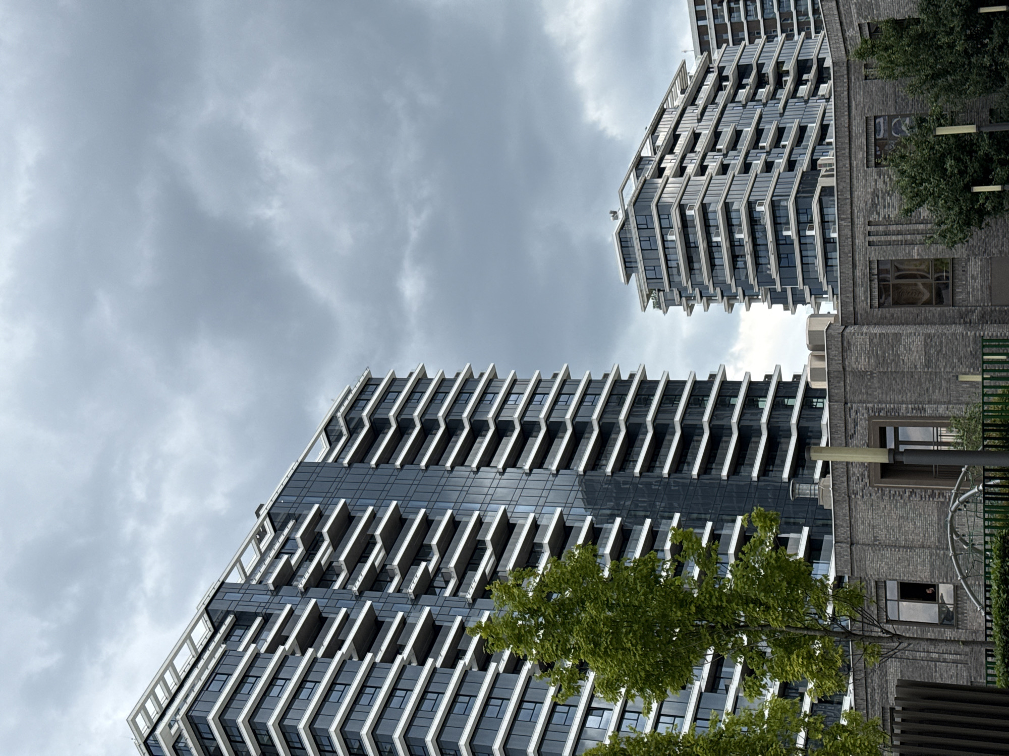 The image shows two modern buildings in Belgrade, Serbia, under a cloudy sky. The building in the foreground has a distinctive facade with alternating rows of windows and white, angled balconies. A green tree partially obscures the lower floors of this building. To the right, another building is visible, connected to a brick structure with windows. The brick building appears older than the modern high-rises. The perspective is from ground level, looking upwards at an angle. The overall mood is somewhat overcast due to the cloud cover.