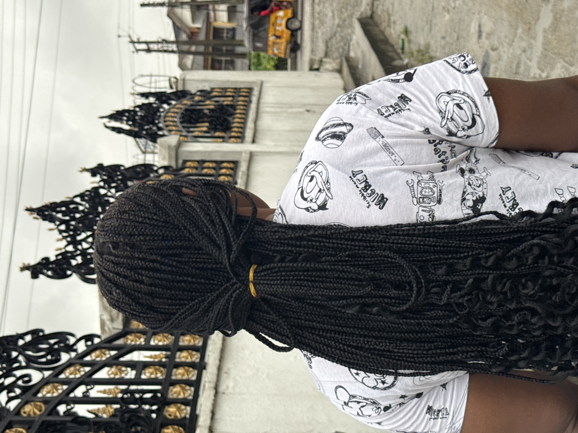 An overhead, angled shot depicts a dark-skinned person with black braided hair, secured by a gold hair tie, in front of a gate made of wrought iron with gold decorative accents. The person is wearing a white top with black cartoon character prints. Behind the gate, there is a white building. In the background, a blurred yellow tricycle is visible. The location is likely Ado, Nigeria, based on the prompt.