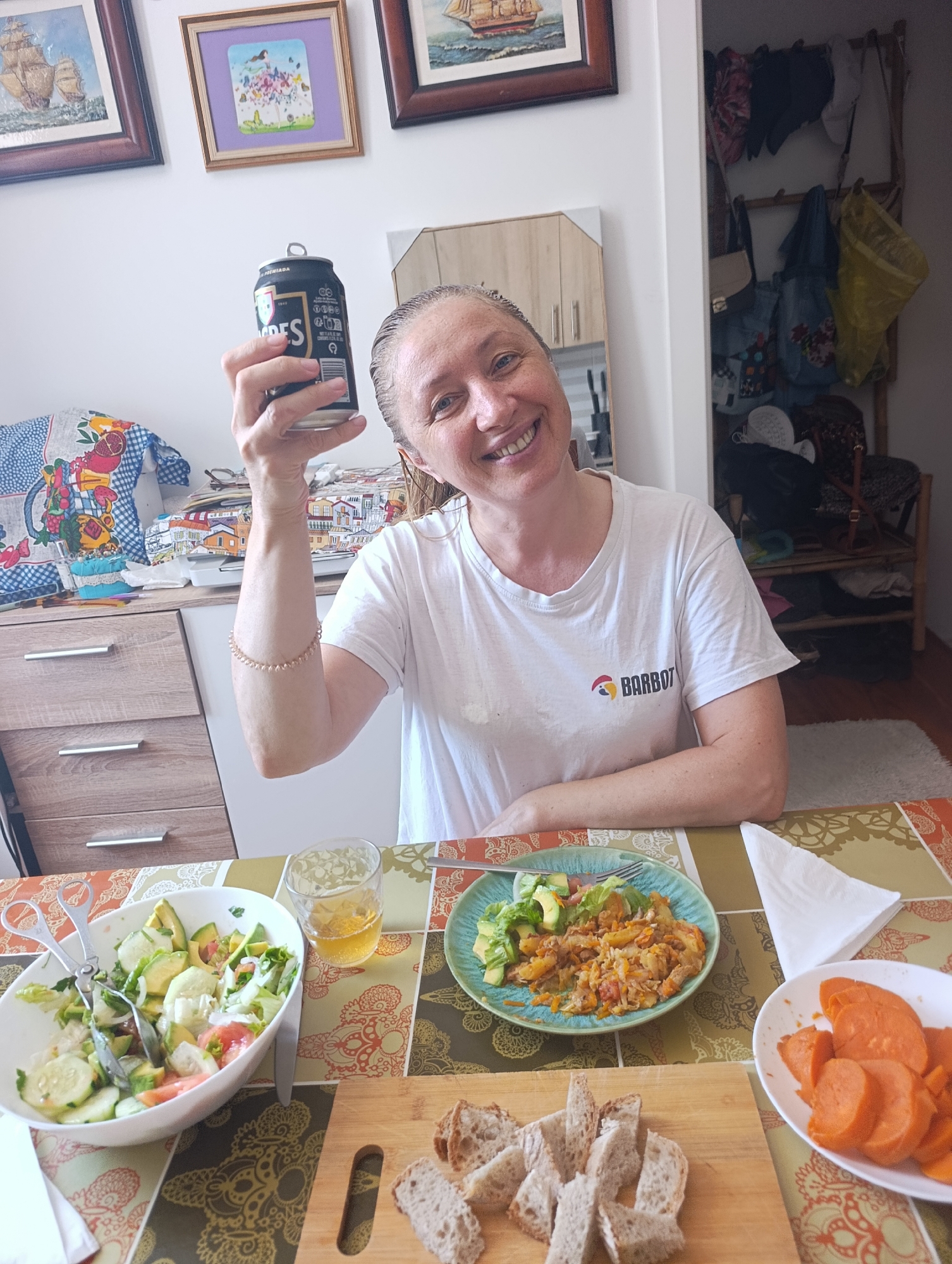 A woman is seated at a table with food and drink. She holds up a can of beer in her right hand, which is positioned to the left of her head. The woman is wearing a white t-shirt with a logo on the left chest that reads "BARBOT." Her left arm is resting on the table, and she is smiling. In front of her is a meal consisting of a salad with lettuce, cucumber, tomato, and avocado in a white bowl with salad tongs. There is also a plate with a cooked dish, likely a stew or stir-fry with vegetables and possibly meat. To the right of the plate is a white napkin. Further to the right is a bowl of sliced sweet potatoes. A glass of beer sits next to the salad bowl. In the foreground, a cutting board holds several slices of bread. The table is covered with a tablecloth that has a colorful, patterned design. In the background, to the left, there are framed pictures on a white wall. To the right, there is a closet or shelf unit with various bags and clothing items hanging. The scene appears to be in an indoor setting, possibly a dining room or kitchen, in the city of Setúbal, Portugal.