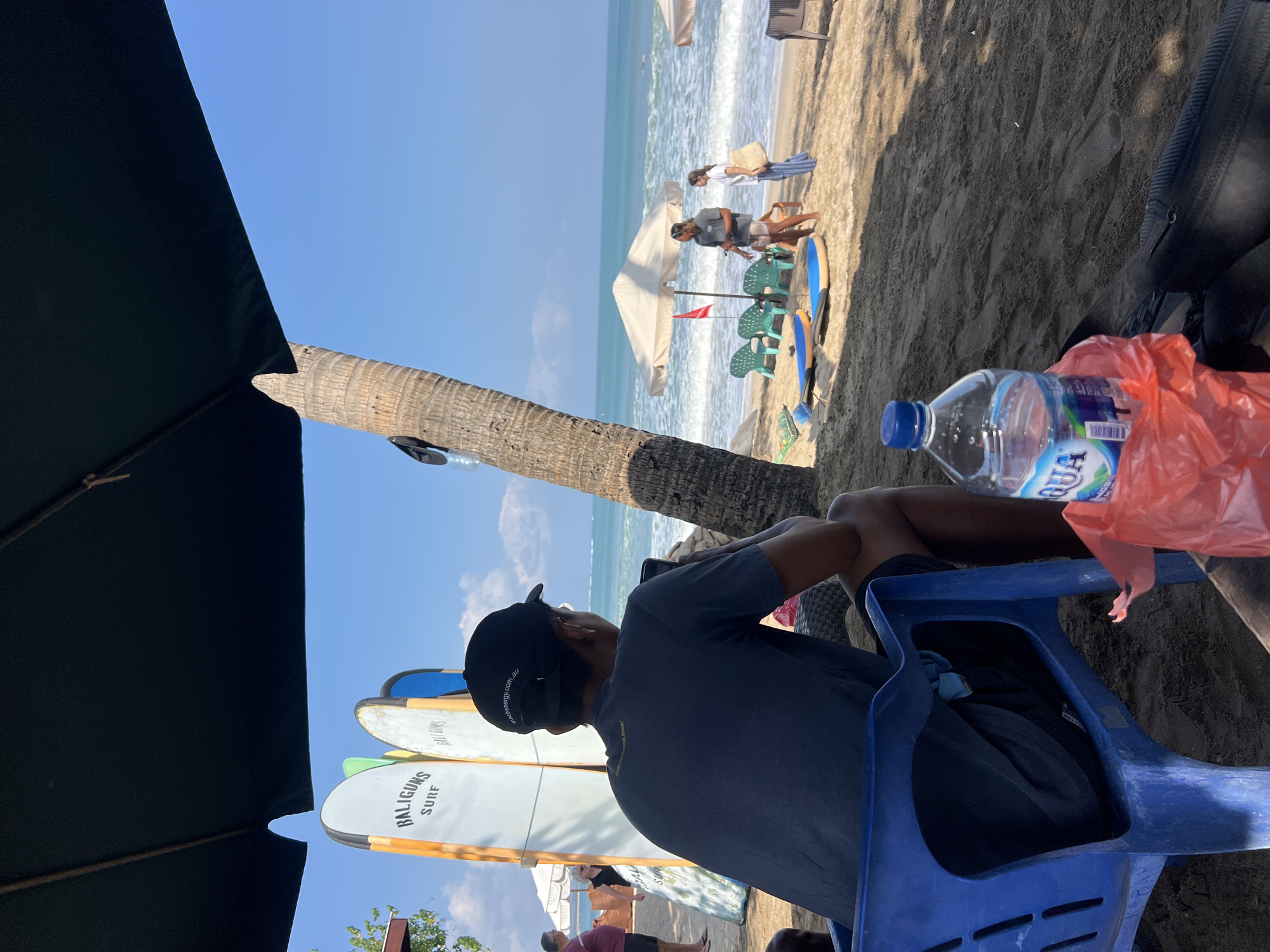 An outdoor, ground-level, medium shot depicts a beach scene in Kuta, Indonesia, on a clear, sunny day. The foreground is dominated by a person sitting in a blue plastic chair, facing away from the camera and looking towards the ocean. This individual wears a dark t-shirt, black shorts, and a black baseball cap. A bottle of Aqua water and a red plastic bag are positioned on the sand in front of the chair.
Behind the seated person, a row of surfboards stands upright, with "BALIGUNS SURF" visible on one. To the right, a palm tree trunk is prominent. The middle ground of the image reveals a beach with people interacting near a large white umbrella and several green plastic chairs. Two surfboards lie on the sand in front of these chairs. The background shows the ocean with waves and a clear blue sky. The scene suggests a relaxed atmosphere at a surf school or beach rental establishment.