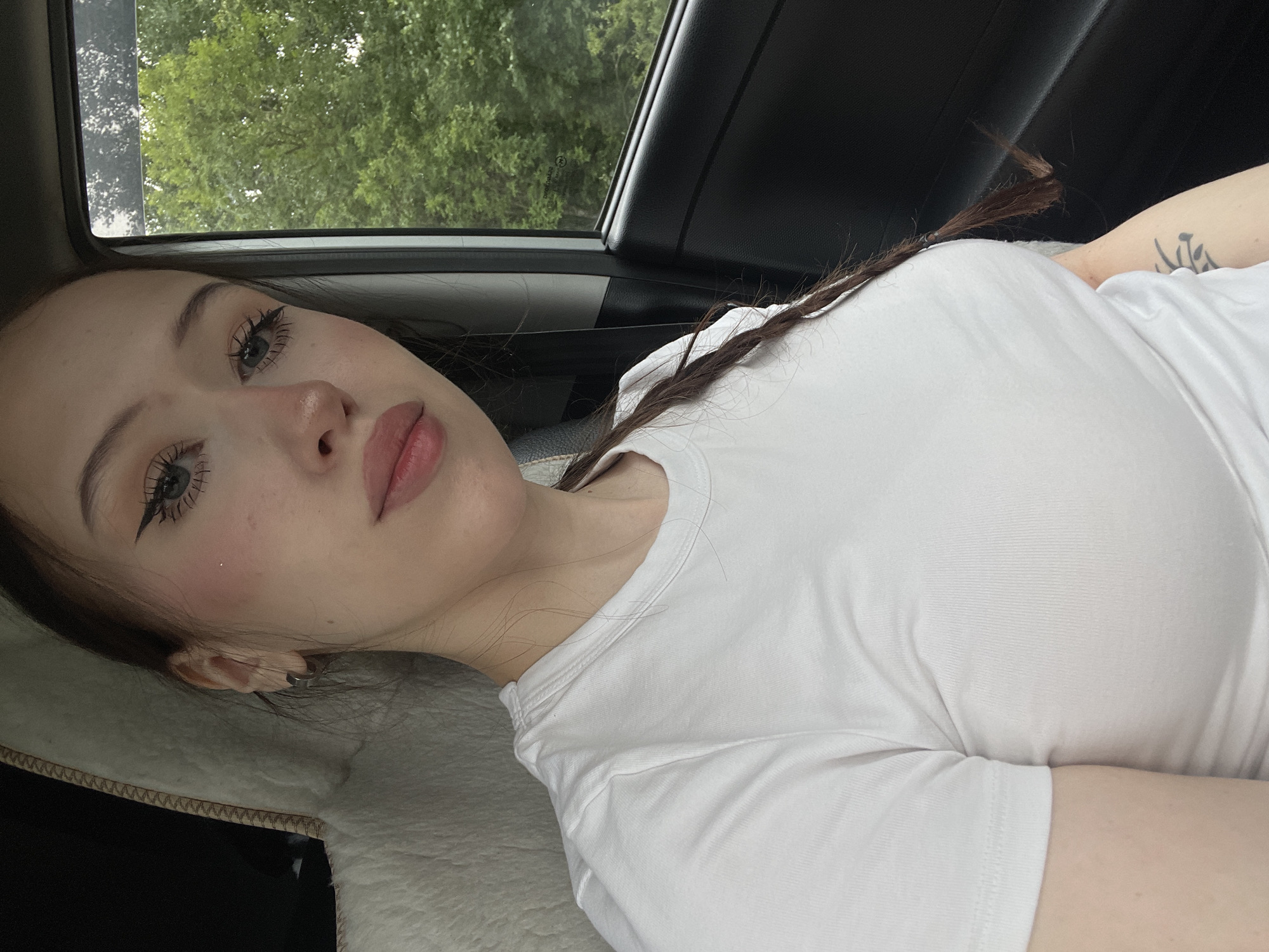 A young woman is captured from the chest up, seated in the passenger seat of a vehicle. She is wearing a white t-shirt and her dark hair is styled in a braid that falls over her shoulder. Her gaze is directed towards the camera, and she has makeup on, including eyeliner and blush. Outside the car window, lush green trees are visible, suggesting a rural or suburban setting. The interior of the car is dimly lit, with the seat fabric visible behind her.