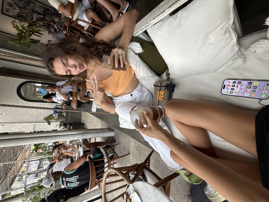 Two women sit outside a café. One woman, angled from the top left, wears a cropped orange top and white pants, holding a beverage, and flashing a peace sign. Her companion, oriented from the bottom right, also holds a beverage. A handbag rests on the cushion between them, and a smartphone is placed on the white cushion of the seating arrangement. Diners are visible at nearby tables.