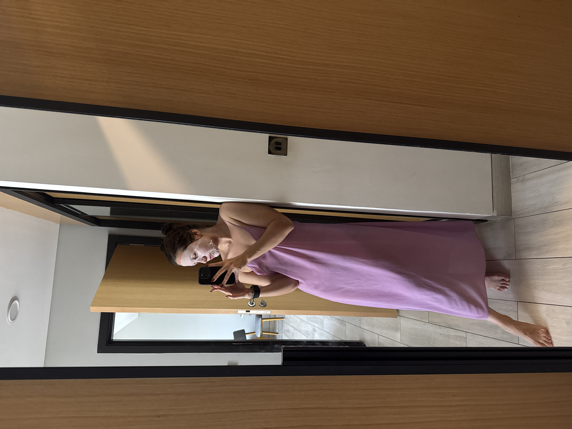 A woman stands in a hotel room, likely in Dubai, United Arab Emirates, based on the modern, minimalist design elements and the style of the architecture visible in the reflection. She is wearing a light purple dress and a facial mask and is taking a picture of herself in a mirror using a smartphone. The room has light-colored walls, wood paneling, and tiled flooring. A black frame surrounds the mirror. There is a partially visible chair in the background through a doorway.