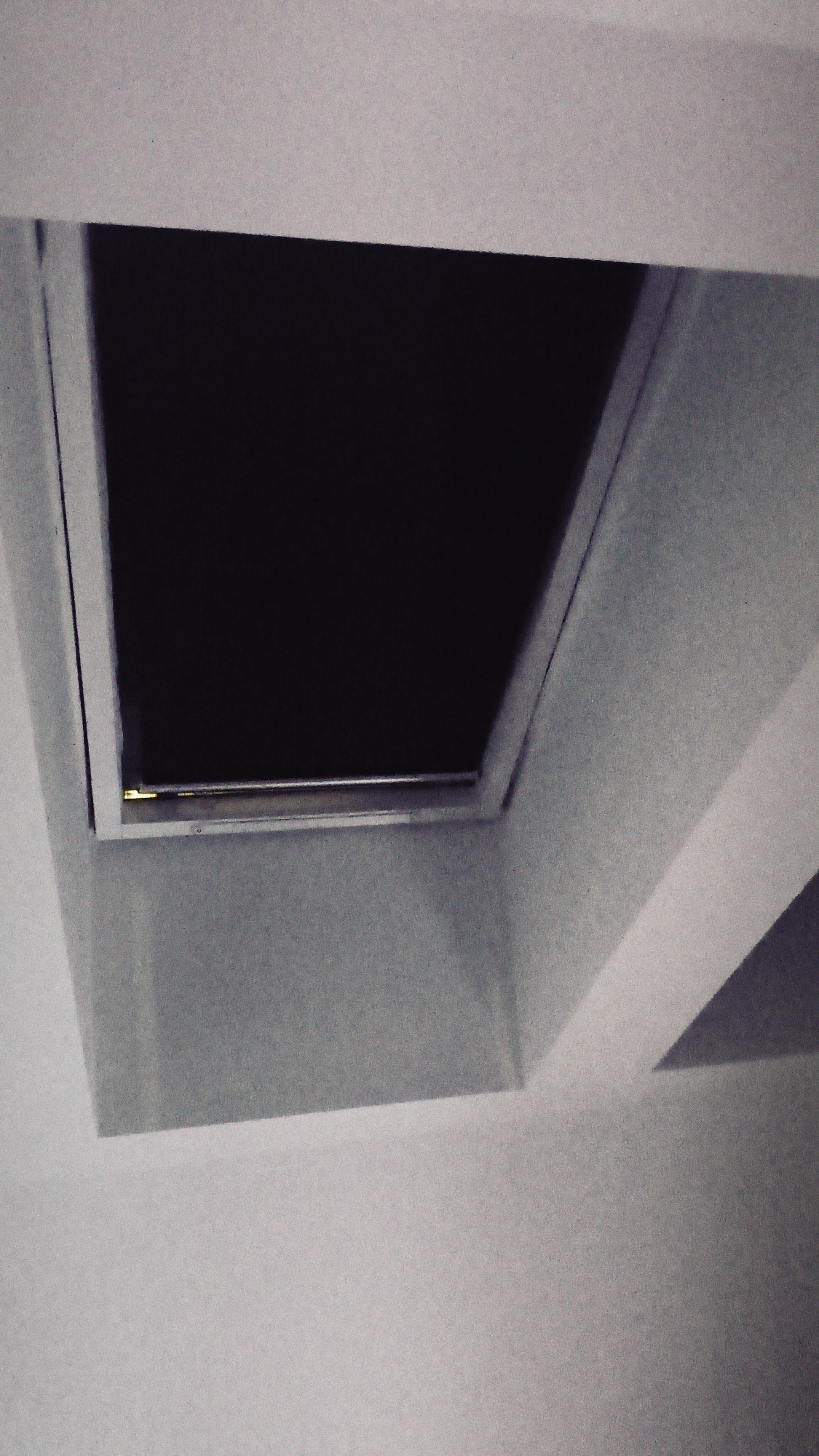 A square skylight is recessed into a white ceiling. The inside of the skylight appears dark, with a metal bar visible across the bottom edge of the opening. Part of another recessed ceiling structure is visible in the lower right corner of the frame. The lighting is dim, and the photograph seems to be taken from a low angle, looking up at the ceiling. It's impossible to determine the specific location in Nowy Tomyśl, Poland, without further information.