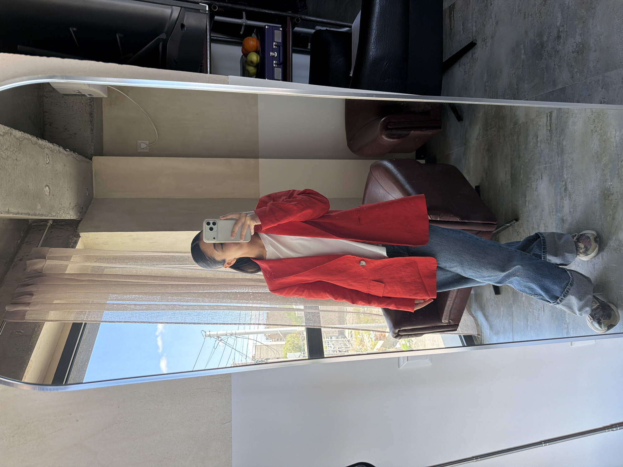 A person stands in front of a full-length mirror, taking a selfie. The person wears a white shirt, blue jeans, and a red blazer. They hold a white smartphone. The reflection in the mirror shows a window with a view of a city street with power lines and buildings. The location is Yerevan, Armenia. The surrounding environment includes a concrete ceiling, neutral-colored walls, and furniture, including a leather chair.