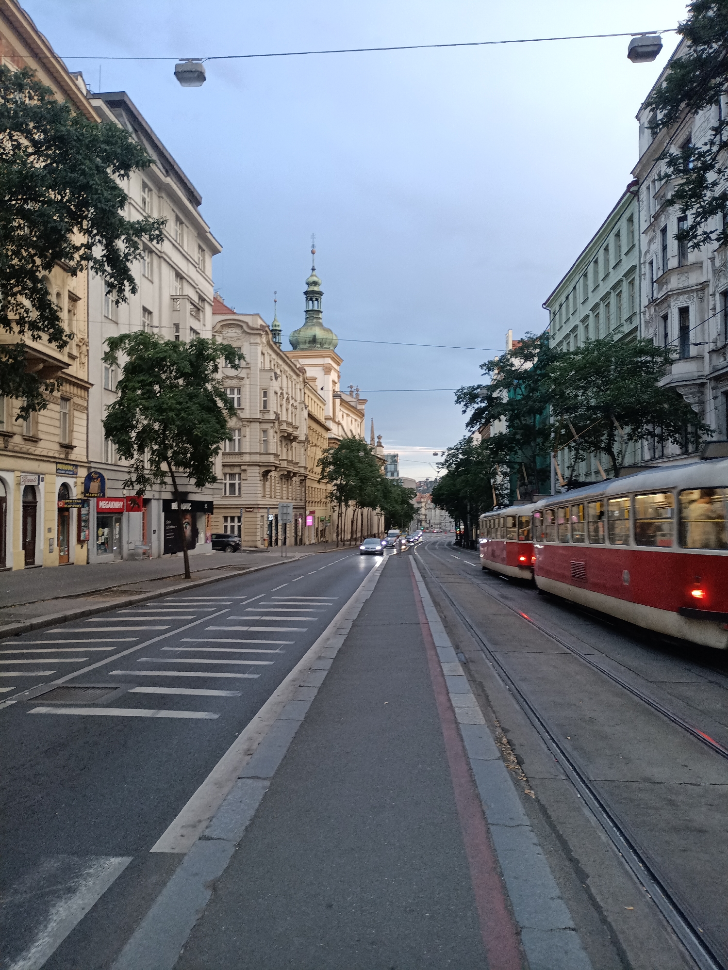 The image depicts a street scene in Prague, Czechia, likely during twilight or overcast conditions, given the soft lighting and muted sky. The perspective is from street level, looking down a wide avenue lined with multi-story buildings on both sides.
On the left side of the street, several buildings are visible, characterized by classical architectural styles with ornate facades, tall windows, and some with decorative elements. Shops and businesses with signage are apparent at street level, including what appears to be a "MegaKnihy" store. Trees with dense green foliage are planted along the sidewalk. A crosswalk is marked on the road, with parallel white stripes.
On the right side of the street, a red and cream-colored tram is in motion, its windows blurred due to the speed. Tram tracks run along the roadway. The buildings on this side are also multi-story and exhibit a mix of architectural styles, some appearing more modern or recently renovated with pastel-colored facades. More trees are present along this side as well.
In the distance, the street curves slightly, and traffic is visible, including cars and at least one other tram further down the line. A distinctive green-domed tower, possibly a church spire or part of a historic building, rises above the rooftops in the middle distance. Overhead, power lines for the tram are strung across the street, with a modern-looking streetlamp suspended from one of them. The overall impression is a typical urban environment in a European city, with functioning public transportation and a blend of historic and contemporary architecture.
