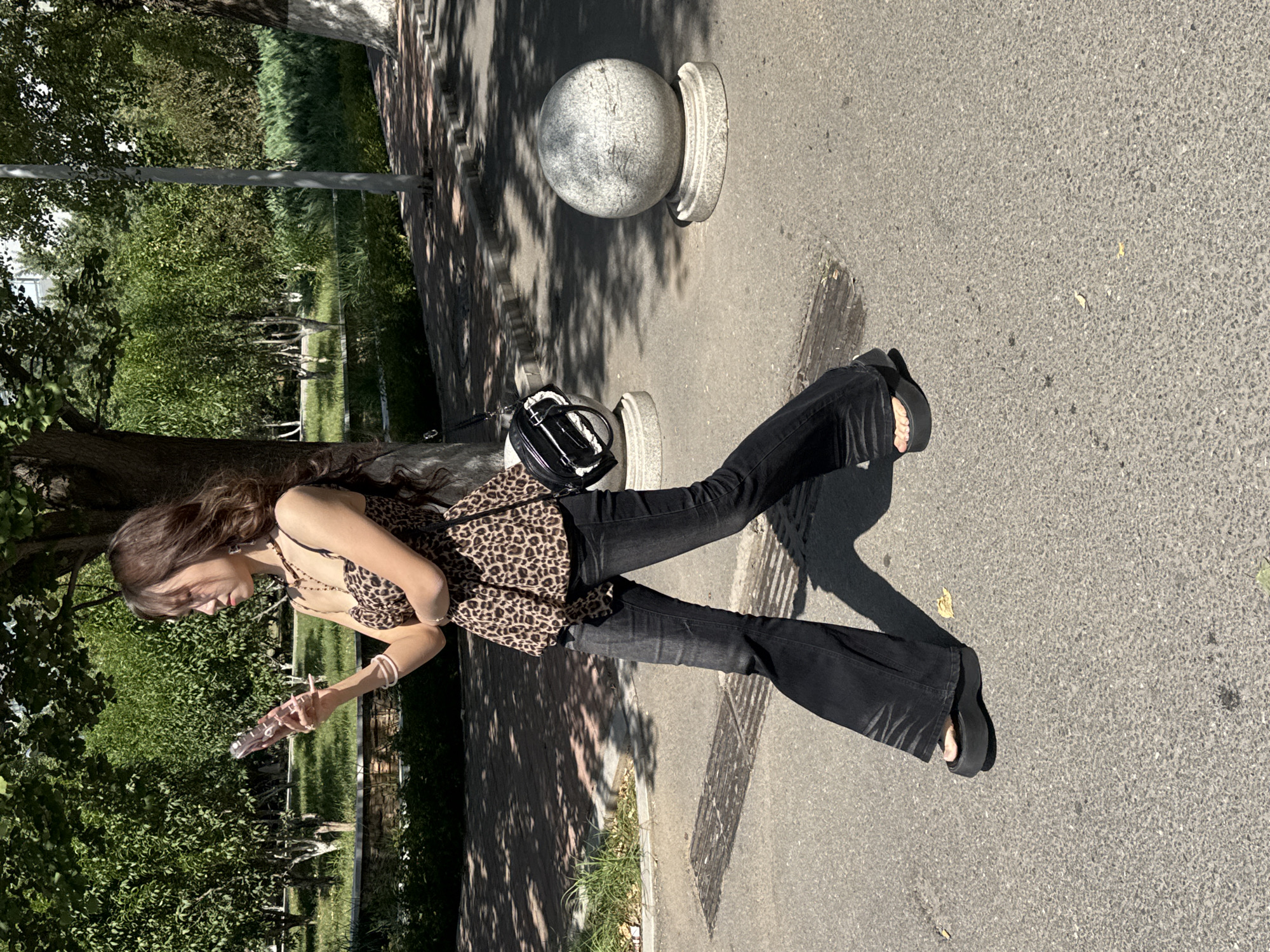 A young woman is walking on a paved sidewalk in Zhongtun, China. She has long, wavy brown hair and is wearing sunglasses. Her outfit consists of a leopard-print halter top, black flared jeans, and black platform sandals. She is holding a pink smartphone in her right hand. A black and white bag with a circular strap is slung over her left shoulder. The background features lush green trees, grass, and a body of water. In the foreground, there are large, round, gray stone bollards on either side of the sidewalk. The lighting suggests it is daytime, with shadows cast by the trees.