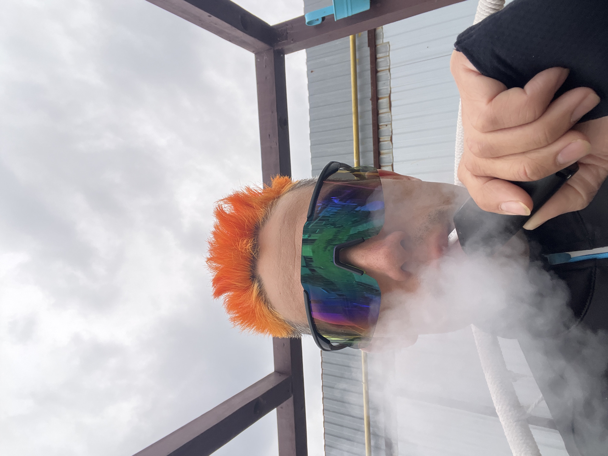 The image features a person's upper body, seen from a low angle, exhaling a large cloud of white vapor. The person has vibrant, spiky orange hair and wears large, mirrored sports sunglasses that reflect green, blue, and purple hues. The sunglasses cover the eyes, obscuring facial expressions, while the vapor conceals the lower part of the face.
The person's right hand is visible in the upper right portion of the frame, holding a dark object, likely a vaping device, close to the mouth area. The fingers are slightly curled, and fingernails are visible.
The background suggests an outdoor setting under a bright, cloudy sky. Dark wooden beams, possibly part of a structural frame, are prominent on the left side of the composition. To the right, a wall of light grey corrugated metal siding is visible, featuring a vertical yellow pipe and a small light blue fixture attached higher up. A thick, white braided rope runs vertically in the mid-ground, positioned to the right of the person. The sky, showing light grey clouds, dominates the upper left and lower portions of the frame. No text is visible in the image.