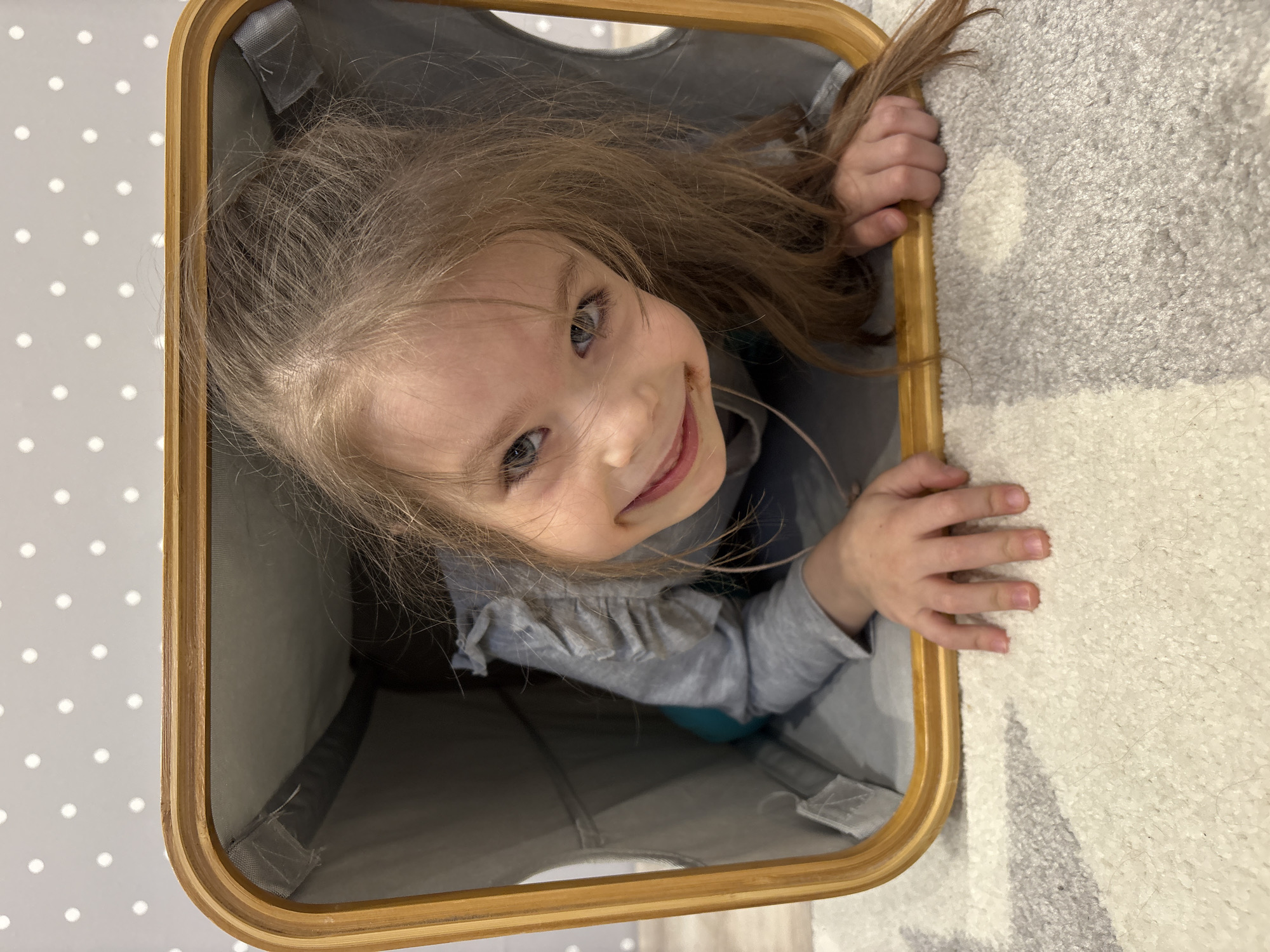 A young child, likely a girl with long, flowing light brown hair and bright blue-gray eyes, is captured in a playful moment, peeking out from a container. She is wearing a light gray long-sleeved top with ruffles on the shoulders. The child's face is central to the frame, beaming with a wide, joyful smile that conveys happiness and amusement. Her eyes are sparkling and focused upwards towards the camera, and her hands are visible gripping the wooden rim of the container. The container itself is lined with gray fabric and features a light wooden top frame. The photo is taken from a top-down perspective, looking into the container, which rests on an indoor floor. To the left, a light gray surface with white polka dots is visible, while to the right, a plush, light-colored carpet with subtle abstract patterns in shades of gray and white can be seen. The indoor lighting suggests a daytime setting. There is no visible text in the image. The scene depicts a heartwarming moment of playful exploration or a game of hide-and-seek. The location context for this domestic scene is Votkinsk, Russia.
