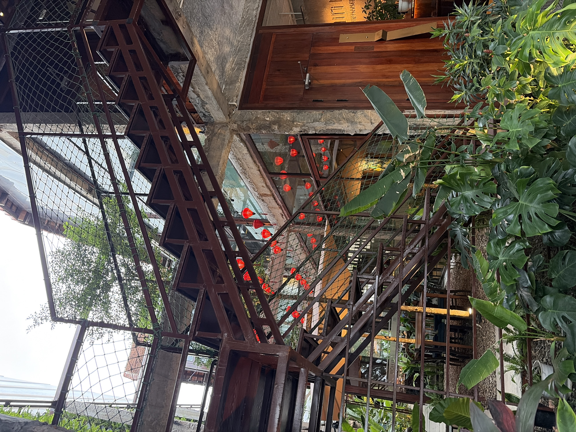 The image depicts the outdoor staircase and surrounding area of a building in Da Nang, Vietnam. The staircase is constructed of dark metal, with open treads and a metal mesh railing. Red paper lanterns are strung along the upper levels, some illuminated. Lush green plants, including large-leafed varieties like Monstera, are abundant at the base of the staircase and along the building's facade. A wooden door is visible on the right side of the image, and large windows reveal interior spaces with seating. The overall aesthetic suggests a restaurant or cafe with an industrial yet tropical design. The lighting indicates it is either late afternoon or early evening.