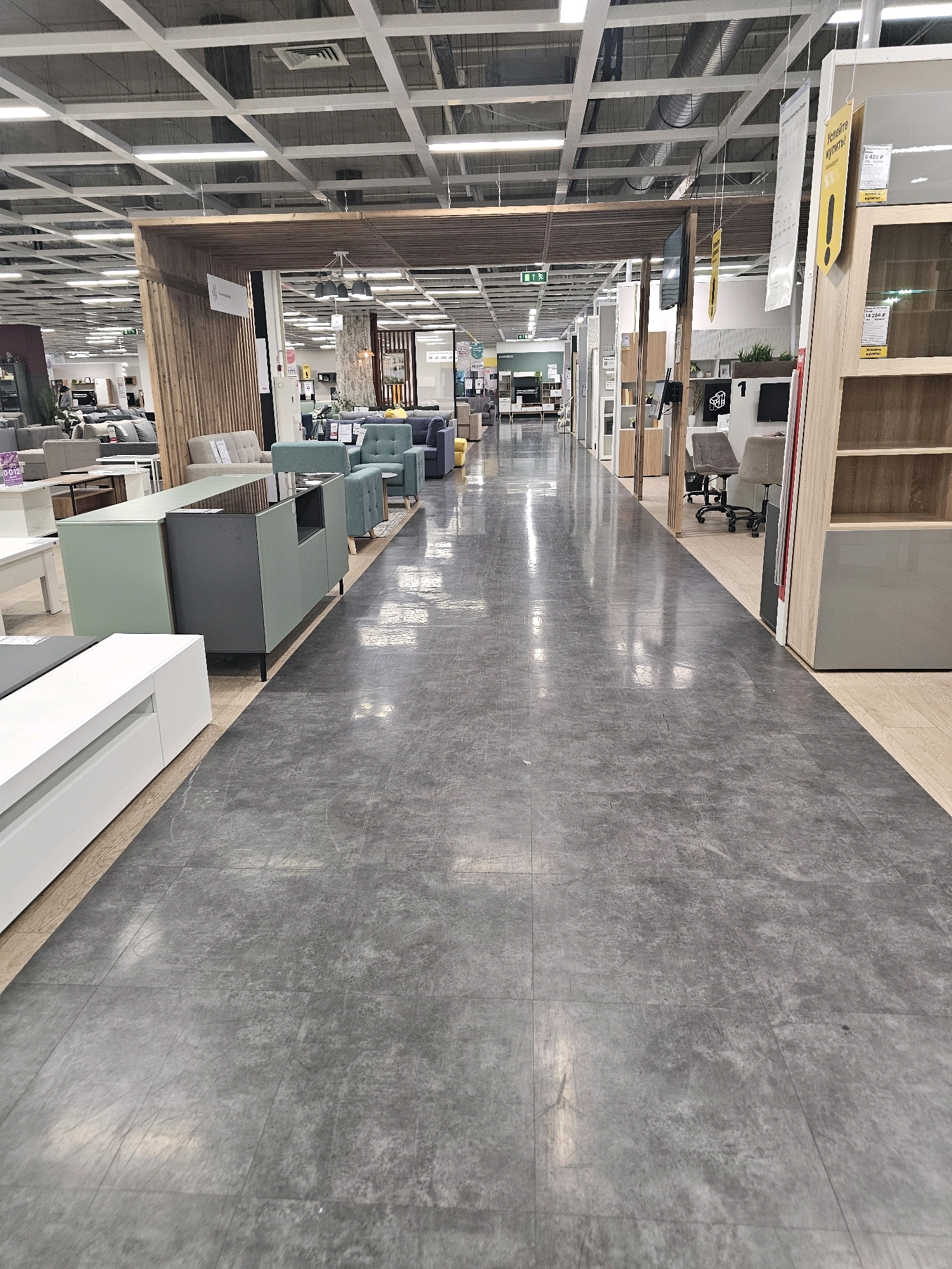 The image shows an interior view of a large furniture store, likely a showroom or retail space, with rows of furniture and shelving visible. The flooring is a dark gray, matte material that appears to be tile or linoleum, laid in large square sections. The ceiling is a suspended grid system with exposed beams, ductwork, and fluorescent lighting fixtures.
In the foreground, a long walkway or aisle extends into the distance. To the left of the aisle, a light-colored media console is partially visible, followed by a dark gray and a light green cabinet or credenza. Further back on the left, a section of a room display includes sofas and armchairs in shades of gray and blue.
On the right side of the aisle, the space opens up to reveal more furniture displays and shelving units. Some of these include glass-fronted cabinets and wooden shelves. Office chairs are visible in a designated area. Yellow and white signage is hung from the ceiling throughout the store.
The overall impression is of a well-lit, modern retail environment designed to showcase furniture. Given the context provided about Moscow, Russia, this scene is likely from a large furniture retailer such as IKEA or a similar establishment within the city. There are no individuals clearly visible or engaged in any significant actions within the frame. The image is static and captured from a stationary viewpoint, looking down the main aisle of the store.