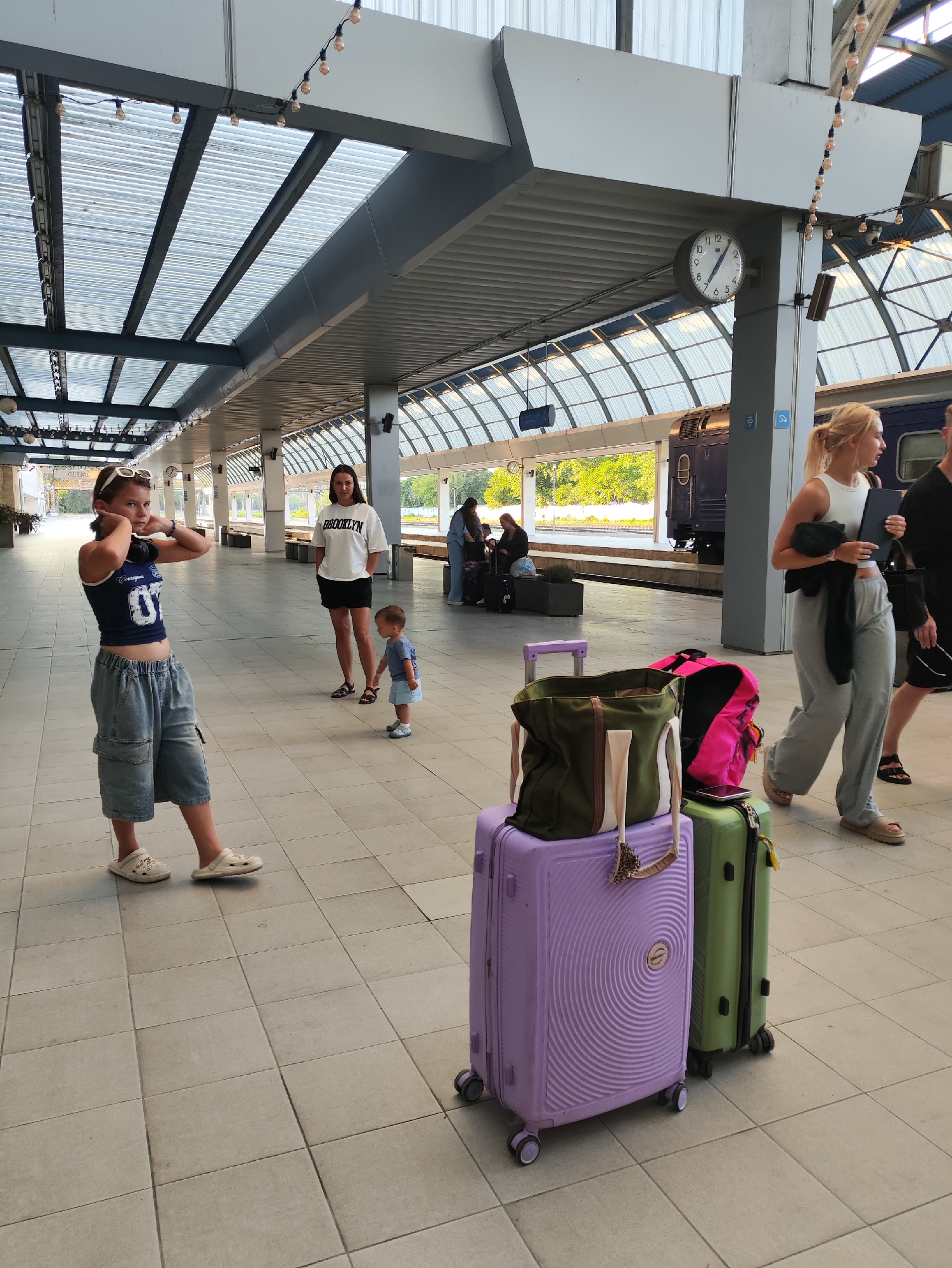 This image captures a scene at a train station, likely in Chișinău, Moldova, given the provided context. In the foreground, a young girl wearing a blue tank top with the number "07" and denim cargo shorts stands with her hands near her ears. Beside her are two suitcases, one large and purple with a distinctive swirl pattern, and a smaller, green suitcase. A large, green canvas bag and a pink backpack rest on top of the purple suitcase.
Further back, a woman in a white t-shirt with "BROOKLYN" printed on it and black shorts stands with her hands in her pockets, looking towards the child. A small child in a blue t-shirt and shorts stands between the two women, holding hands with the woman in the "BROOKLYN" shirt.
In the background, other individuals and luggage are visible, and a train with a dark blue carriage is parked at the platform. The station features a translucent roof supported by a modern architectural structure. A large clock is mounted on a pillar, showing approximately 1:55. The floor is tiled with square gray tiles. The overall lighting suggests it is daytime.