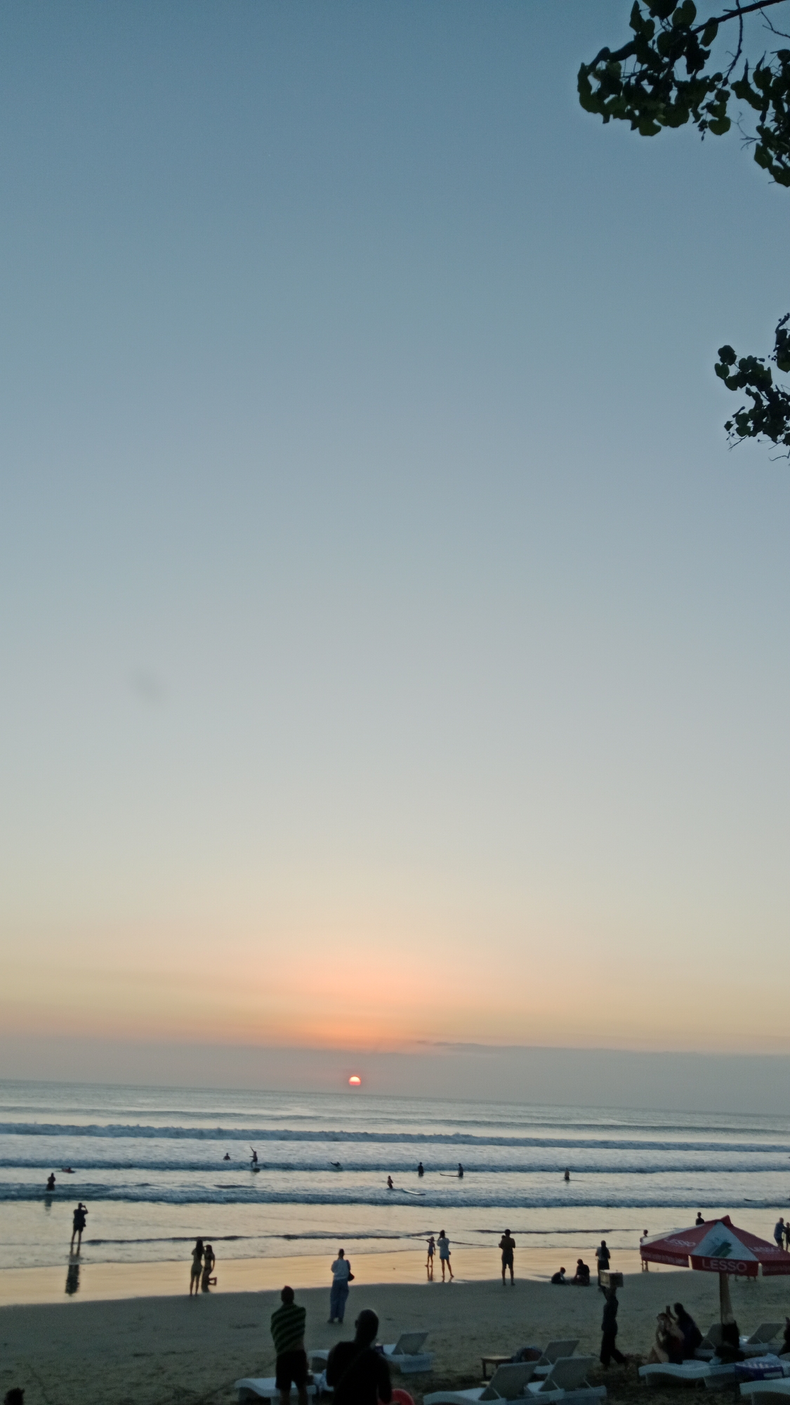 The image depicts a beach scene in Kuta, Indonesia, at sunset. The sky is a gradient of soft blue and peach, with the sun a small, reddish-orange orb low on the horizon. The ocean has gentle waves, and several people are visible in the water, some with surfboards, suggesting they are surfing or enjoying the water. The beach is sandy, with a scattering of people walking, standing, or sitting on lounge chairs. There are also beach umbrellas, one of which is red and white with the word "LESSO" visible. Some people appear to be taking photos or observing the sunset. The overall atmosphere is calm and relaxed, characteristic of a beach at dusk.