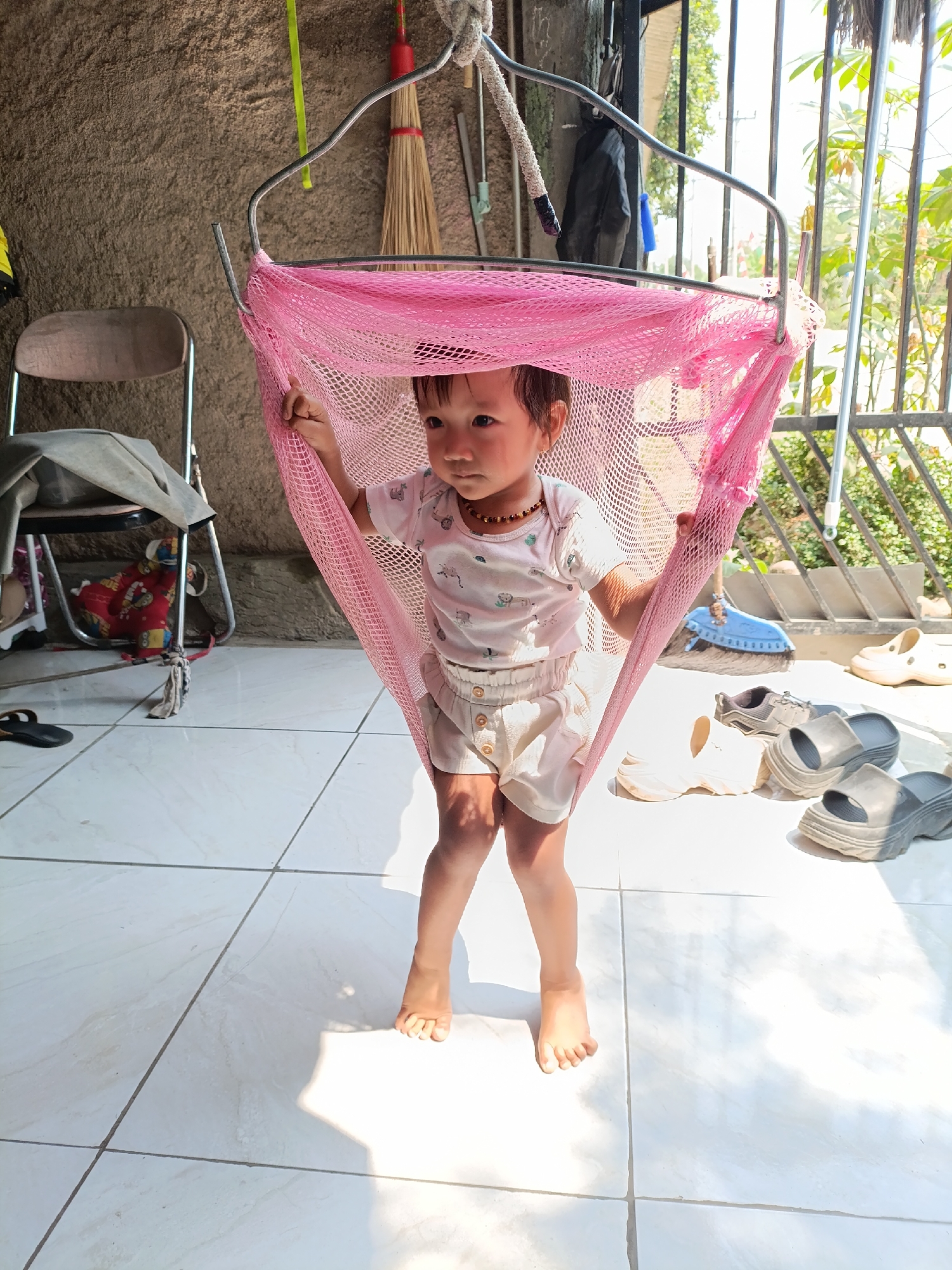 A young child stands in a pink net hammock, supported by a metal frame. The child wears a white t-shirt with animal prints and light-colored shorts. The hammock is suspended from a metal structure, possibly a ceiling or beam. The background reveals a tiled floor, a concrete wall, a chair with clothing draped over it, and several pairs of shoes scattered on the floor. In the distance, there is a glimpse of outdoor greenery and fencing. The lighting suggests it is daytime, with sunlight casting shadows on the floor. The scene appears to be an outdoor or semi-outdoor area, potentially a courtyard or porch, within a residential setting in Cariu, Indonesia.