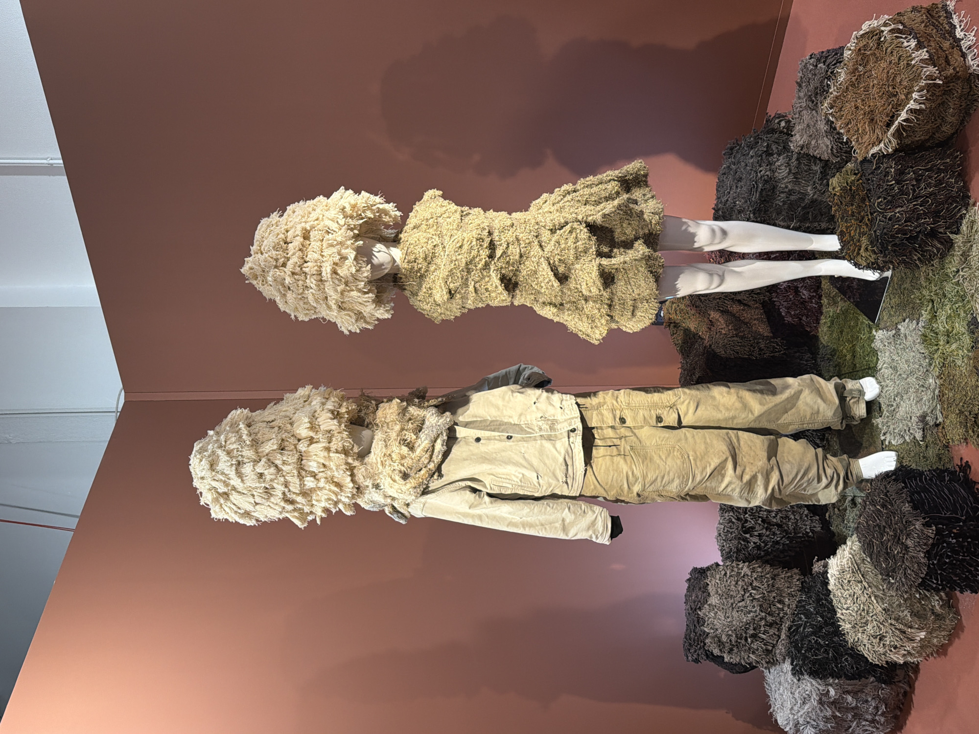 This media file presents an indoor display featuring two mannequins and a collection of textured objects. The background is a solid, warm reddish-brown wall, with a segment of a white ceiling and corner visible in the upper left.
On the left, a female mannequin is dressed in a short, light beige or pale green dress crafted from a shaggy, looped fabric, accompanied by a voluminous, matching headpiece that obscures its head. The mannequin's white legs are visible.
To the right, a male mannequin wears a light beige jacket and light brown cargo pants. It also features a similar shaggy headpiece that covers its head. The mannequin's white feet are visible at the bottom of the pants.
Around the base of the mannequins and spread across the floor to the right are numerous rounded, irregularly shaped objects or cushions. These items share the same distinctive shaggy or tufted texture as the mannequins' attire and headpieces, but are presented in a range of earthy colors including various browns, dark greys, muted greens, and black. They are arranged in a dense pile, forming a textured ground element for the display.
The scene suggests an artistic exhibit or a retail presentation, emphasizing rich textures and an organic, earthy aesthetic through its materials and color scheme. No text is visible in the image.